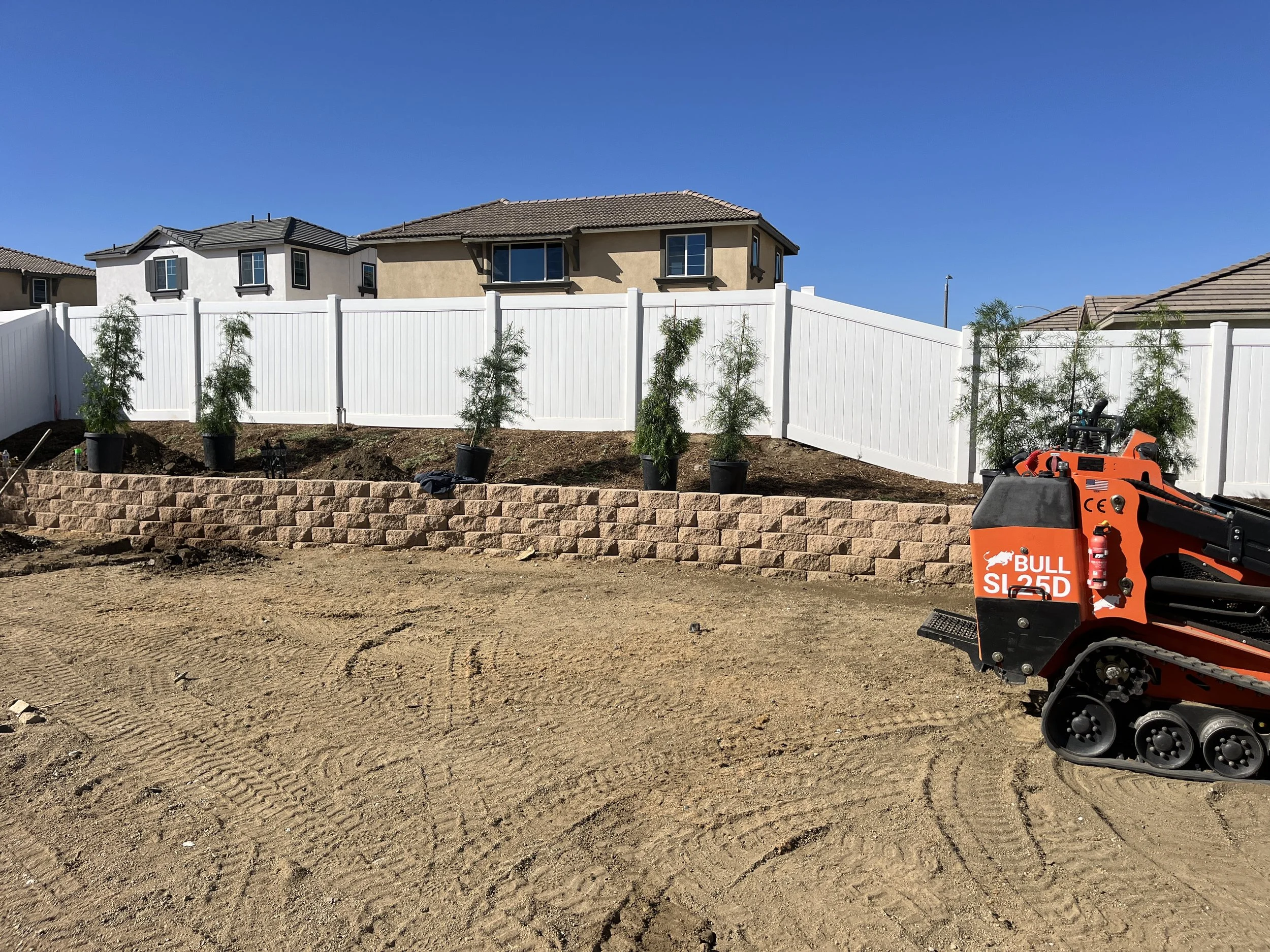 Backyard under construction with a retaining wall of interlocking bricks, potted trees along the wall, and an orange tracked skid steer loader on the dirt ground.