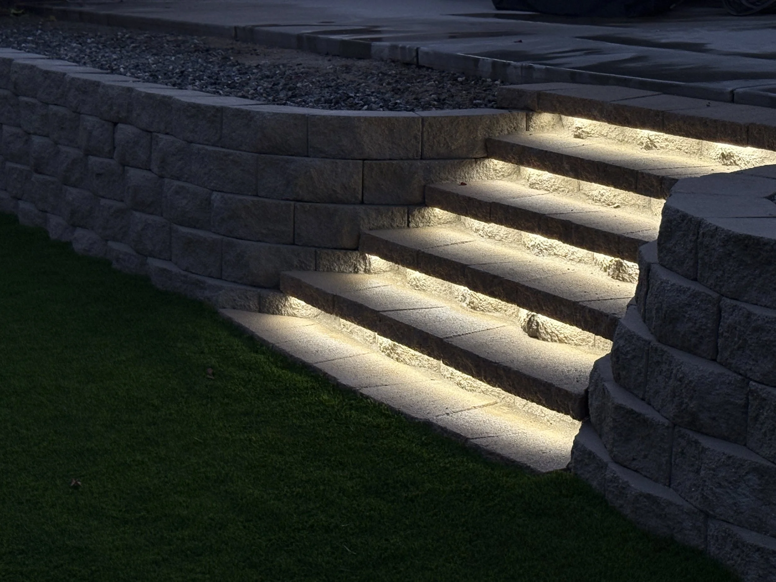 Nighttime outdoor scene of stone steps illuminated by built-in lights, with a grass lawn in the foreground. Backyard landscape renovation in Canyon Lake with locking stone steps, retaining wall, turf, and privacy hedges.