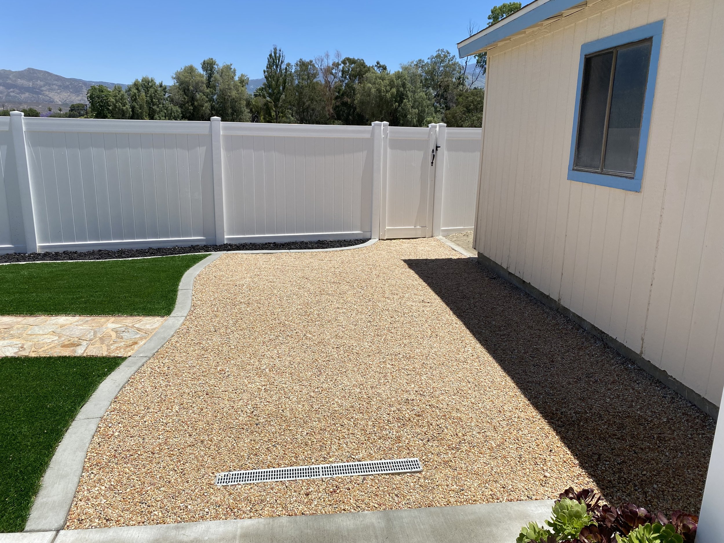 Backyard patio with artificial grass, a gravel area, a white fence with a gate, and a beige house with a window with blue trim.