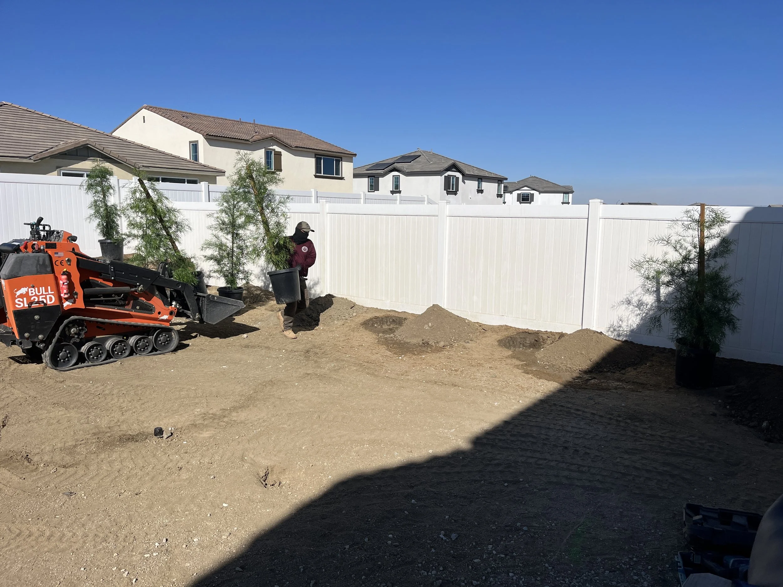 A backyard under construction with a small tracked excavator, a person holding a bucket, and three potted plants against a white fence, with a clear blue sky overhead.