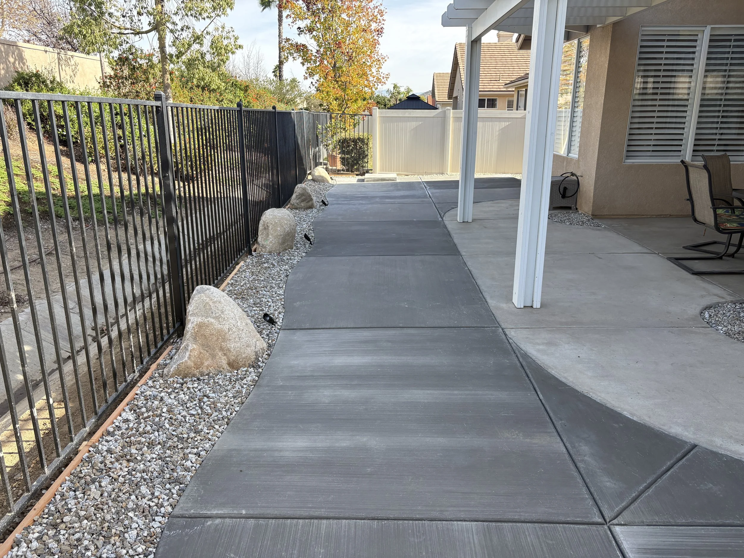A backyard patio with concrete and stamped concrete walkways bordered by gravel, rocks, and a black metal fence. There are a few outdoor chairs along the house wall, and a white support pole on the patio. Residential outdoor upgrade in Murrieta