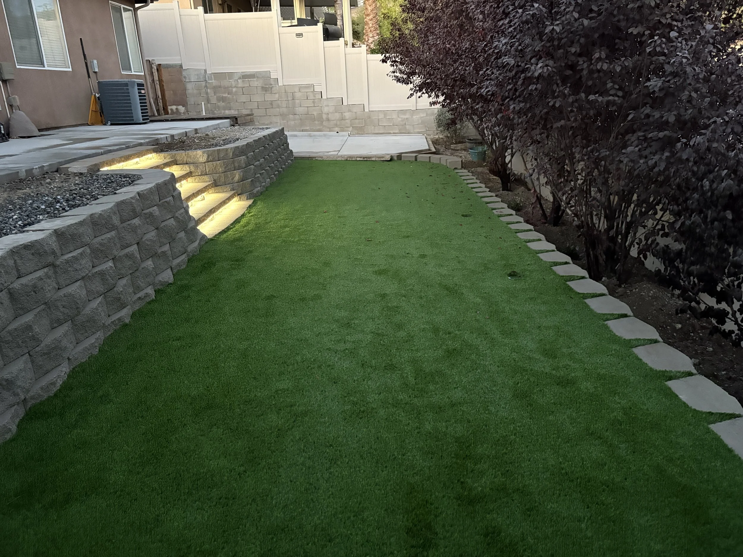 Backyard with manicured artificial grass, stone steps with lighting on the left, a retaining wall with a patio, and a row of dark-leaved bushes on the right. Purple plums bush privacy hedge installation in Canyon Lake, California.
