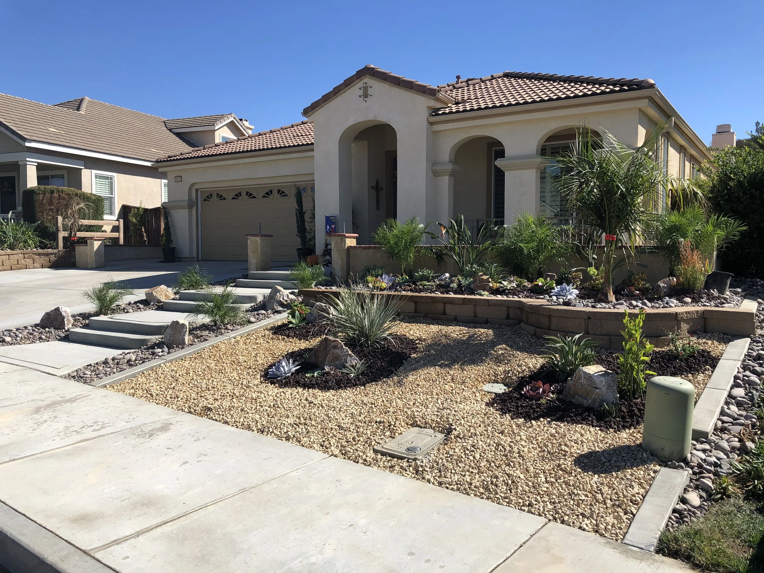 Front yard of a house with desert landscaping, gravel, succulents, and palm trees, with a concrete driveway and stairway leading to the front door.