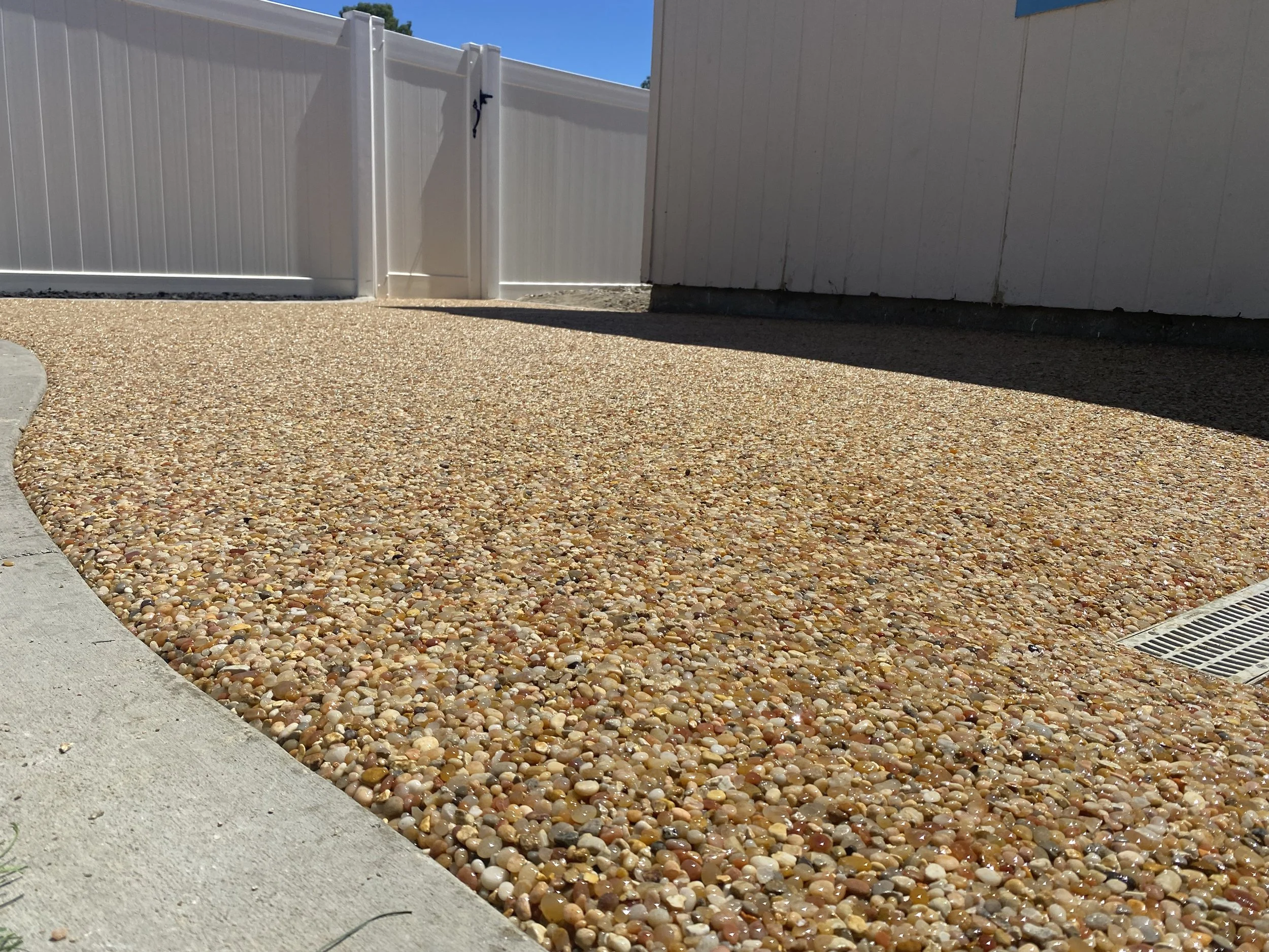 A gravel backyard with a curved concrete border and a white fence with gate, under a clear blue sky.