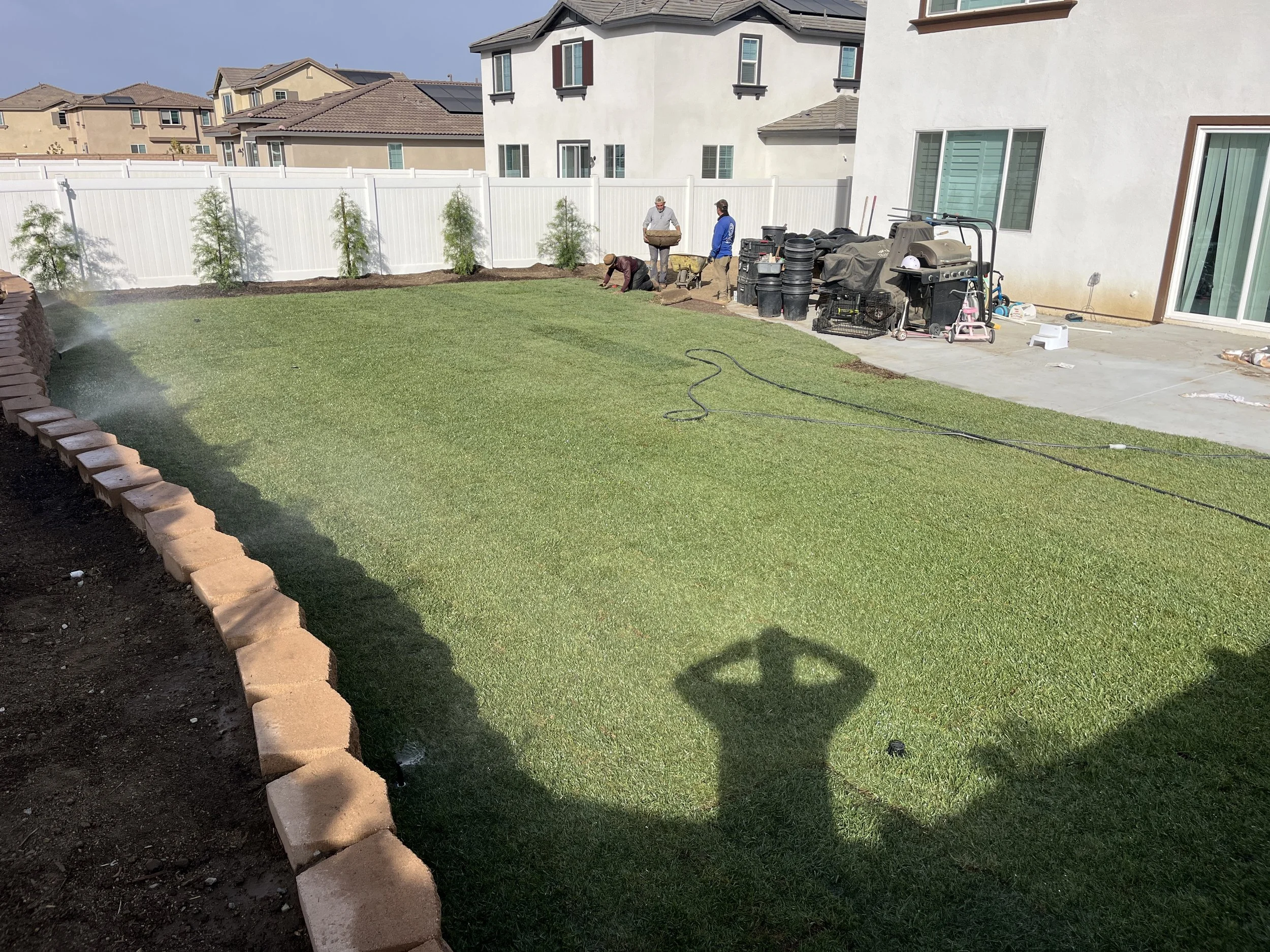 A backyard lawn being landscaped with workers planting plants and preparing the soil along the edge of the grass. Tools and equipment are on the patio, and a white fence surrounds the yard.