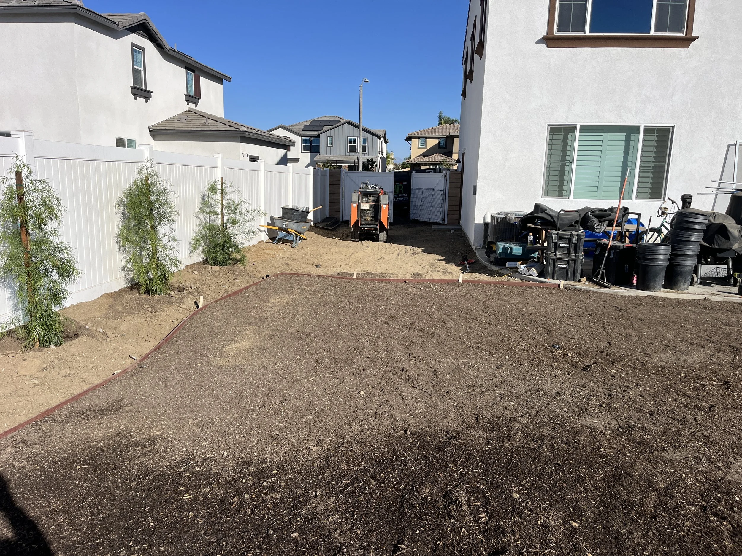 Backyard under construction with dirt and gravel area, small trees along a white fence, construction tools, and building materials, with neighboring houses in the background.