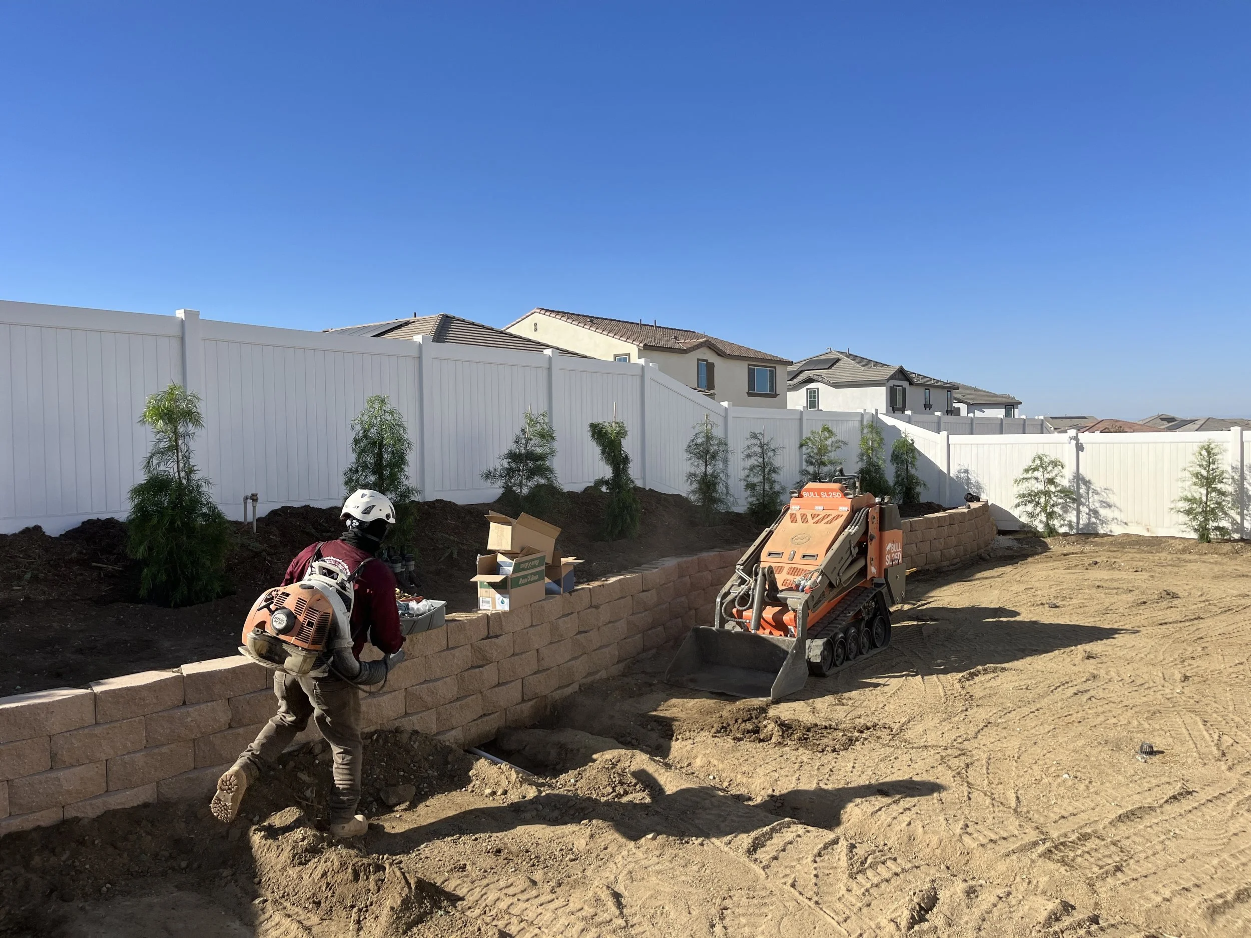 Construction worker operating a small tracked skid-steer loader and building a brick retaining wall in a backyard with a white fence and small trees.
