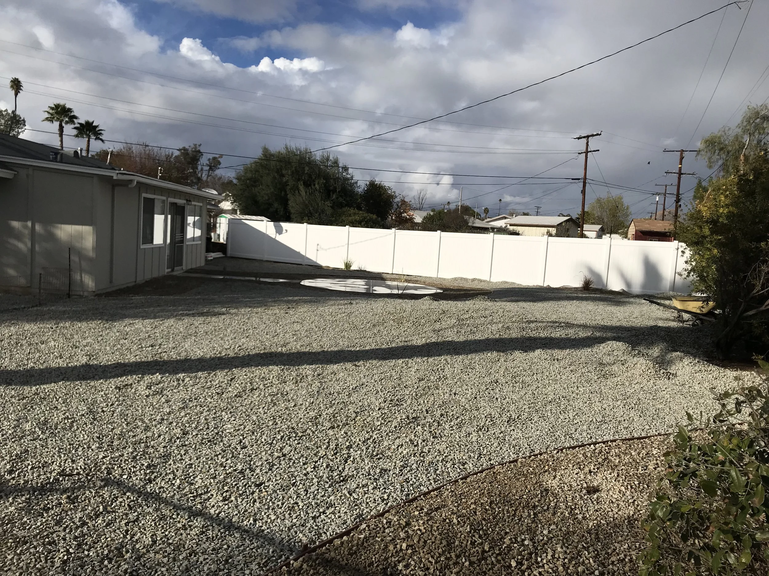 Backyard with gravel ground, a small shed on the left, a white privacy fence in the middle background, a wheelbarrow on the right, and power lines overhead under partly cloudy skies.