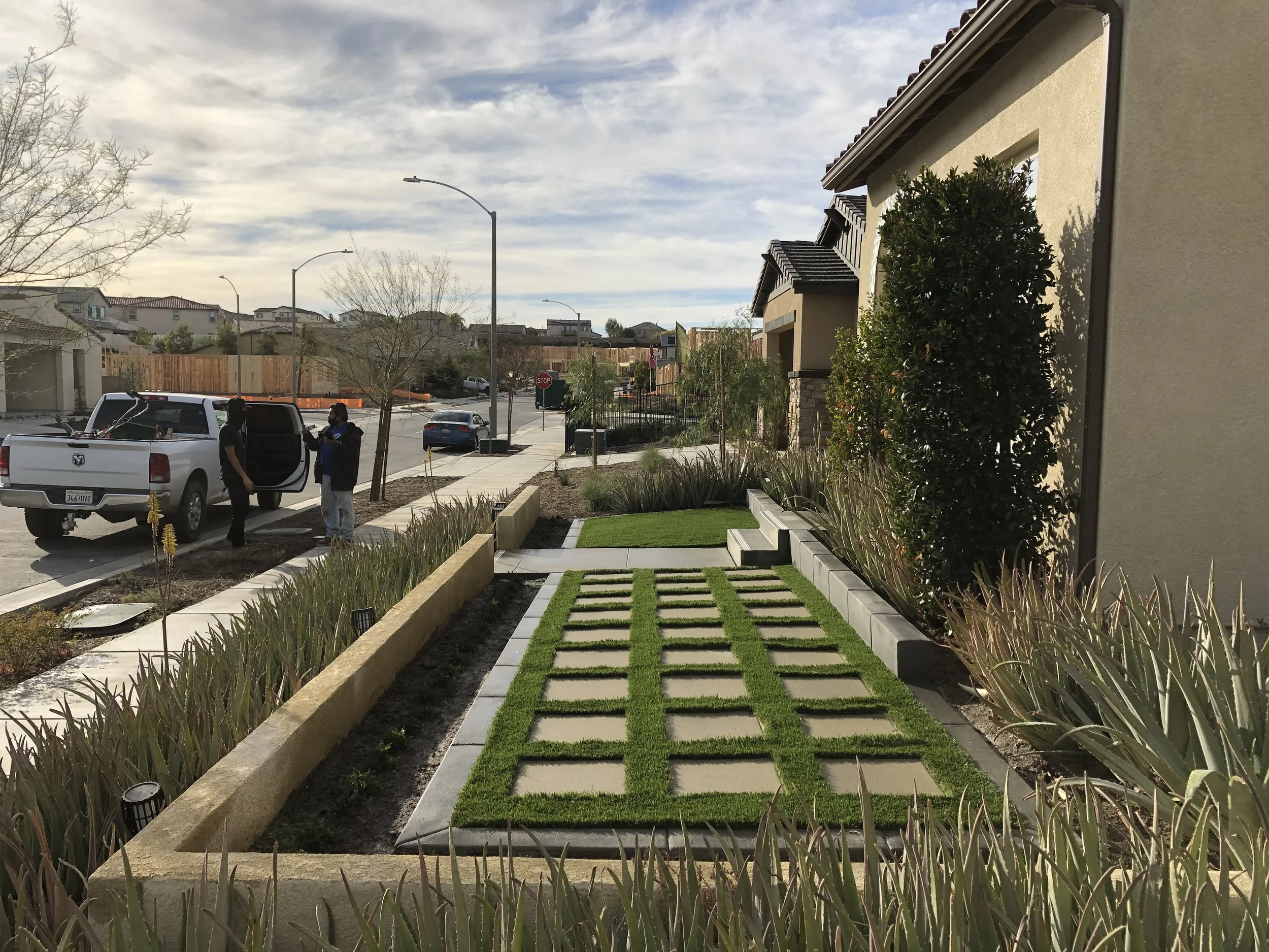 View of a modern residential front yard with a patterned grass and concrete walkway, landscaped plants, and a sidewalk with two people near a white pickup truck, across the street with houses and a stop sign under a partly cloudy sky.