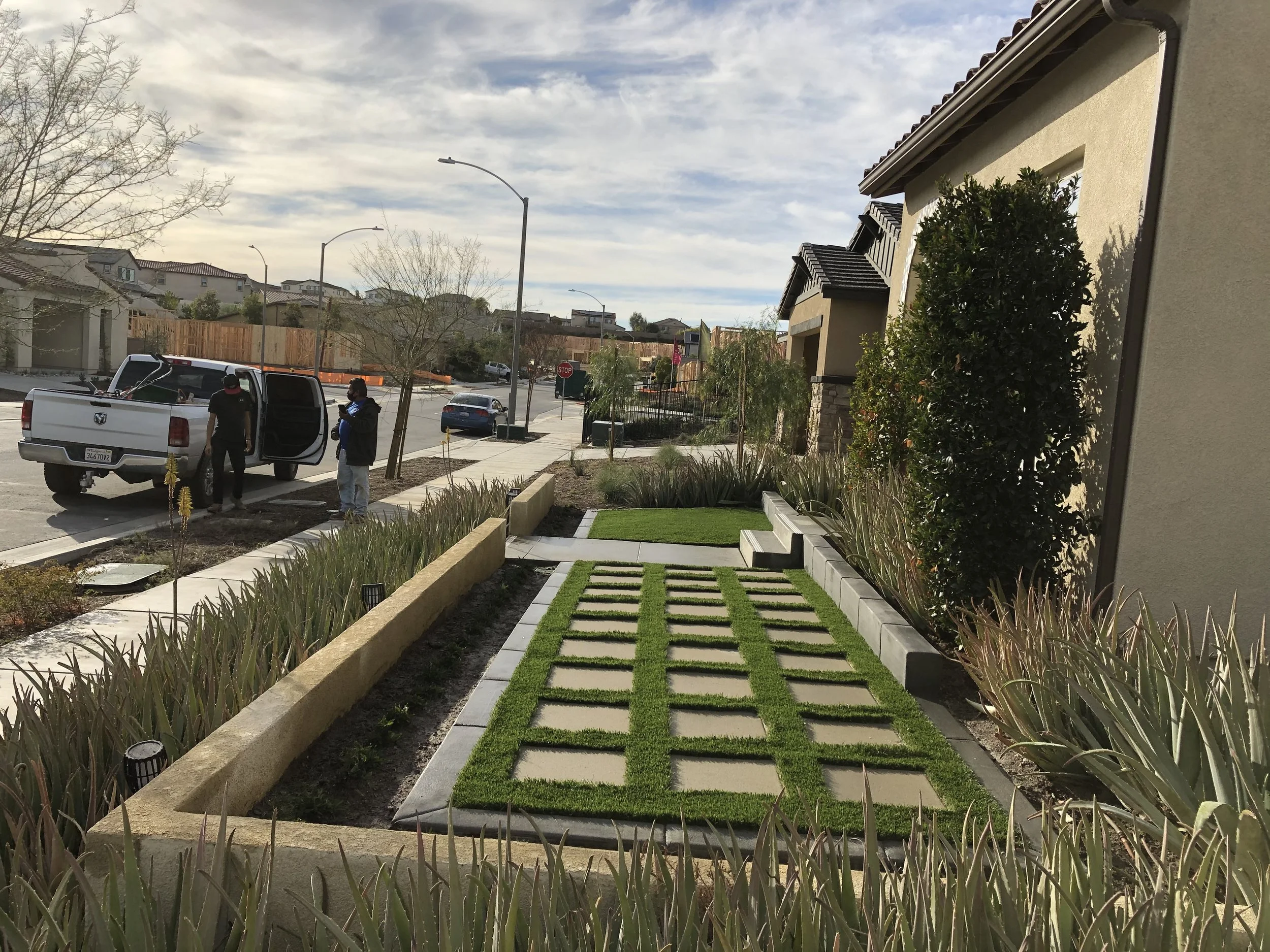 A residential street scene with two men near a white pickup truck, one opening the truck door and another standing nearby. A few cars are parked along the street. The house in the foreground has a small front yard with a tiled pathway, patches of art