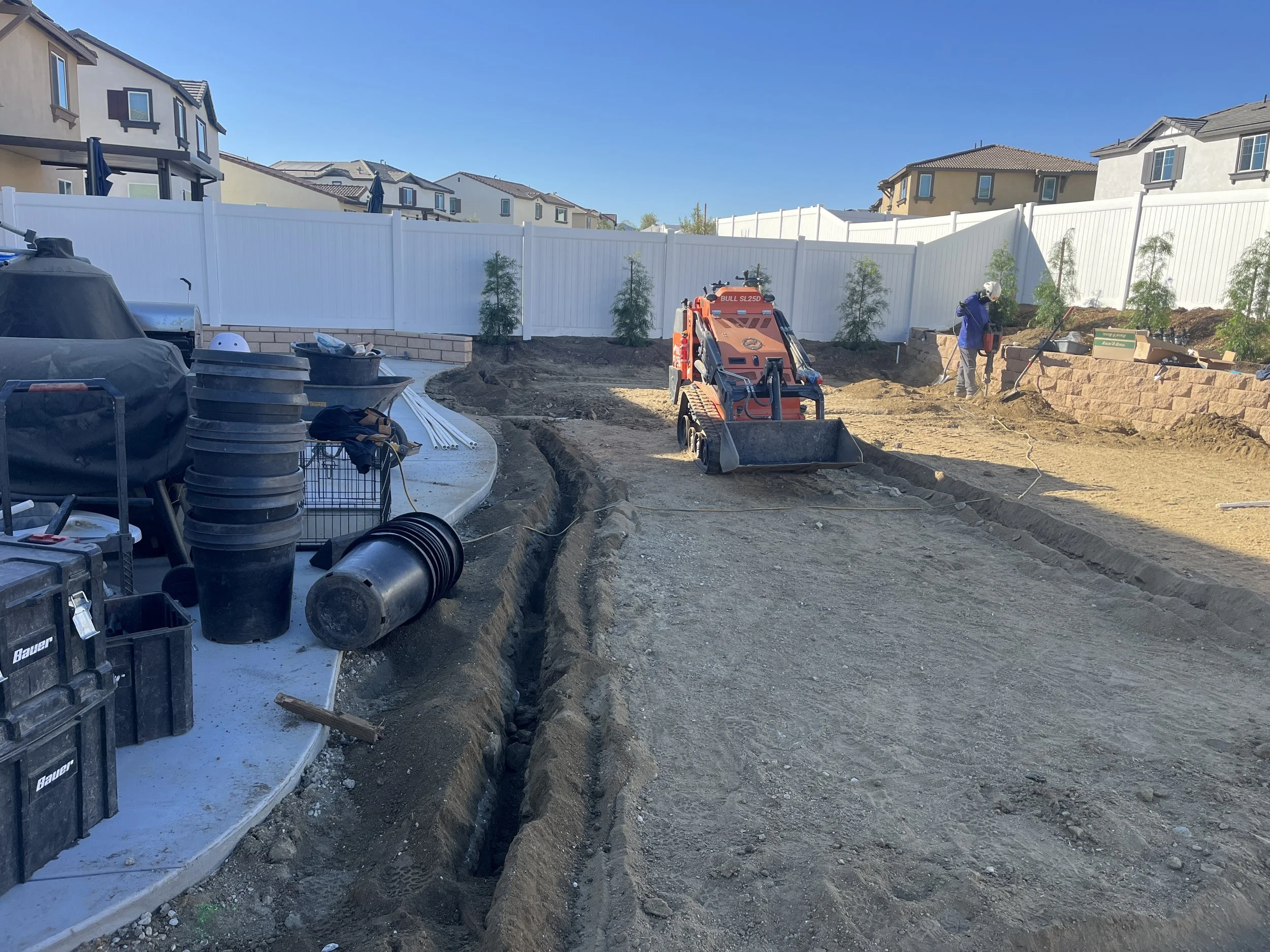 Construction site with a small orange excavator, a worker with a white helmet, and freshly dug trenches beside a row of residential houses with a white fence in the background.
