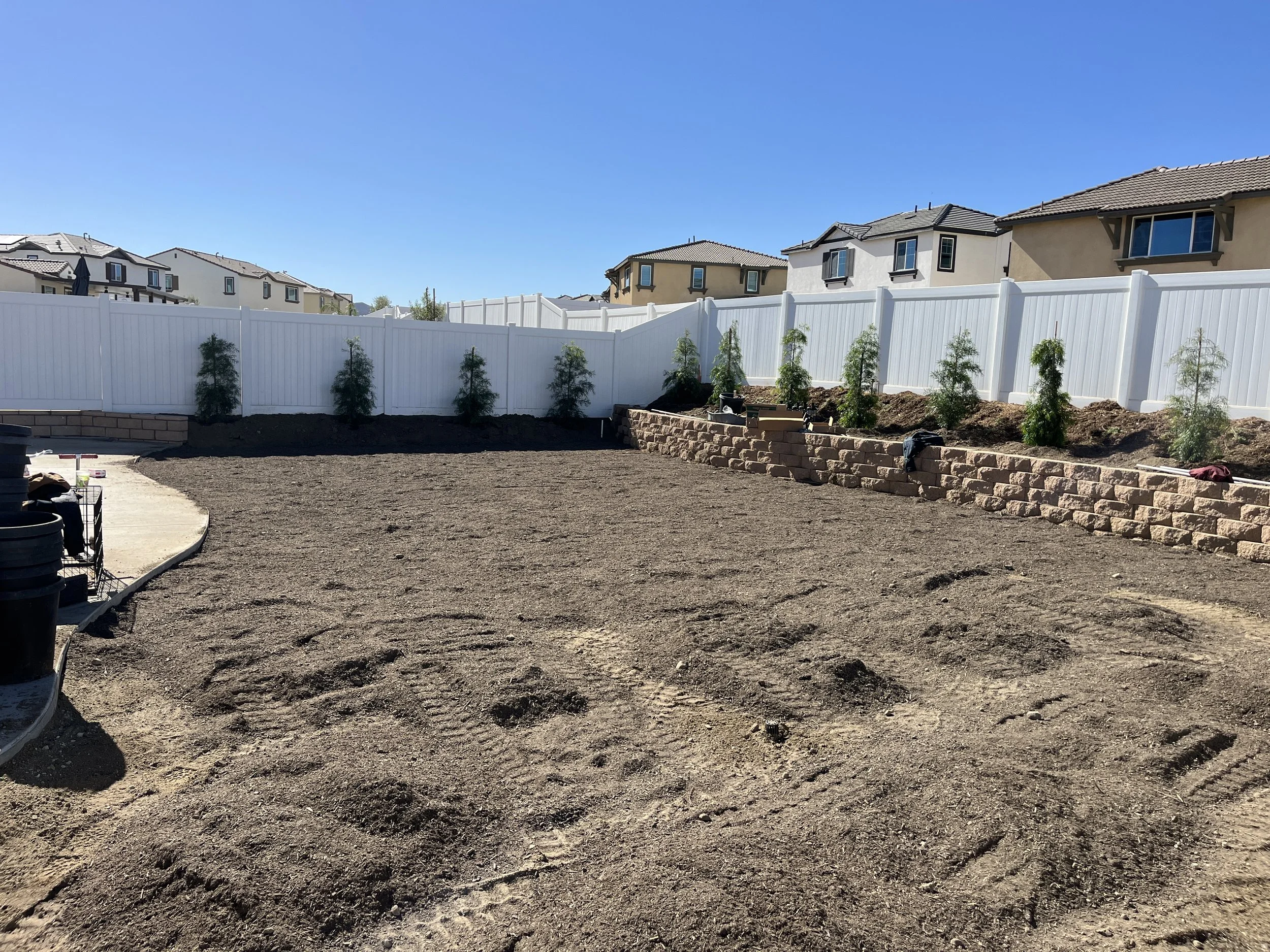 Backyard with a dirt ground, newly planted small trees behind a white fence, and a low brick wall at the far end. Some gardening tools and supplies are visible on the side.