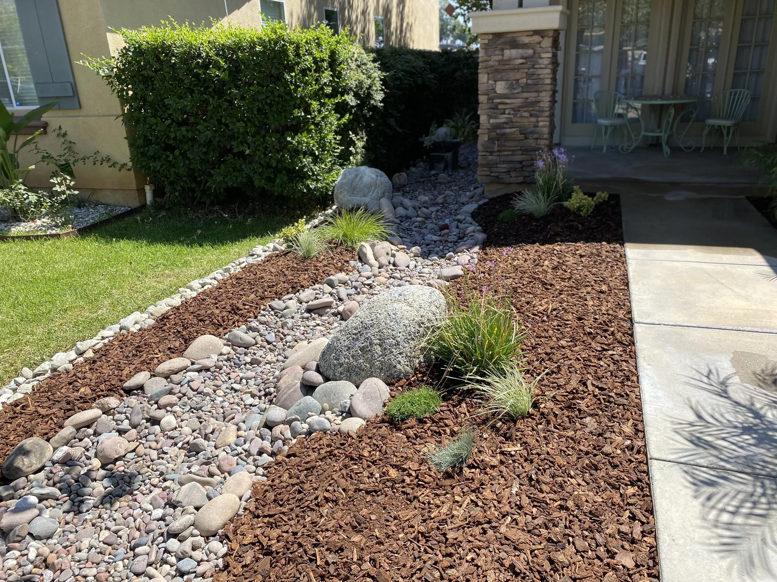 A landscaped yard with a variety of plants, rocks, and mulch, adjacent to a concrete walkway and house with patio furniture.