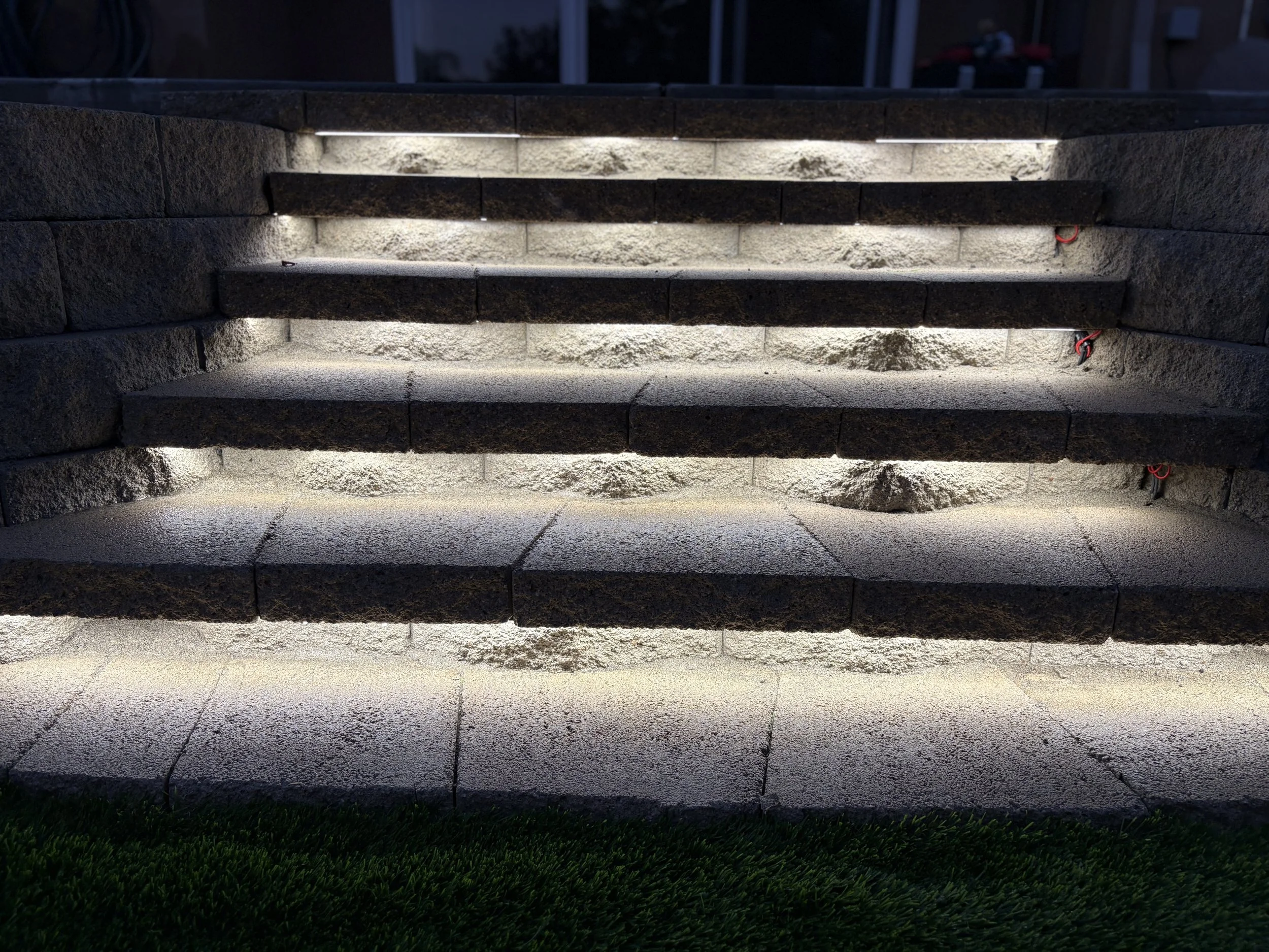 Steps made of dark stone with integrated lighting illuminating the edges, leading upwards, with grass at the bottom and building structures in the background. High-quality artificial turf installation detail in Canyon Lake landscape project.