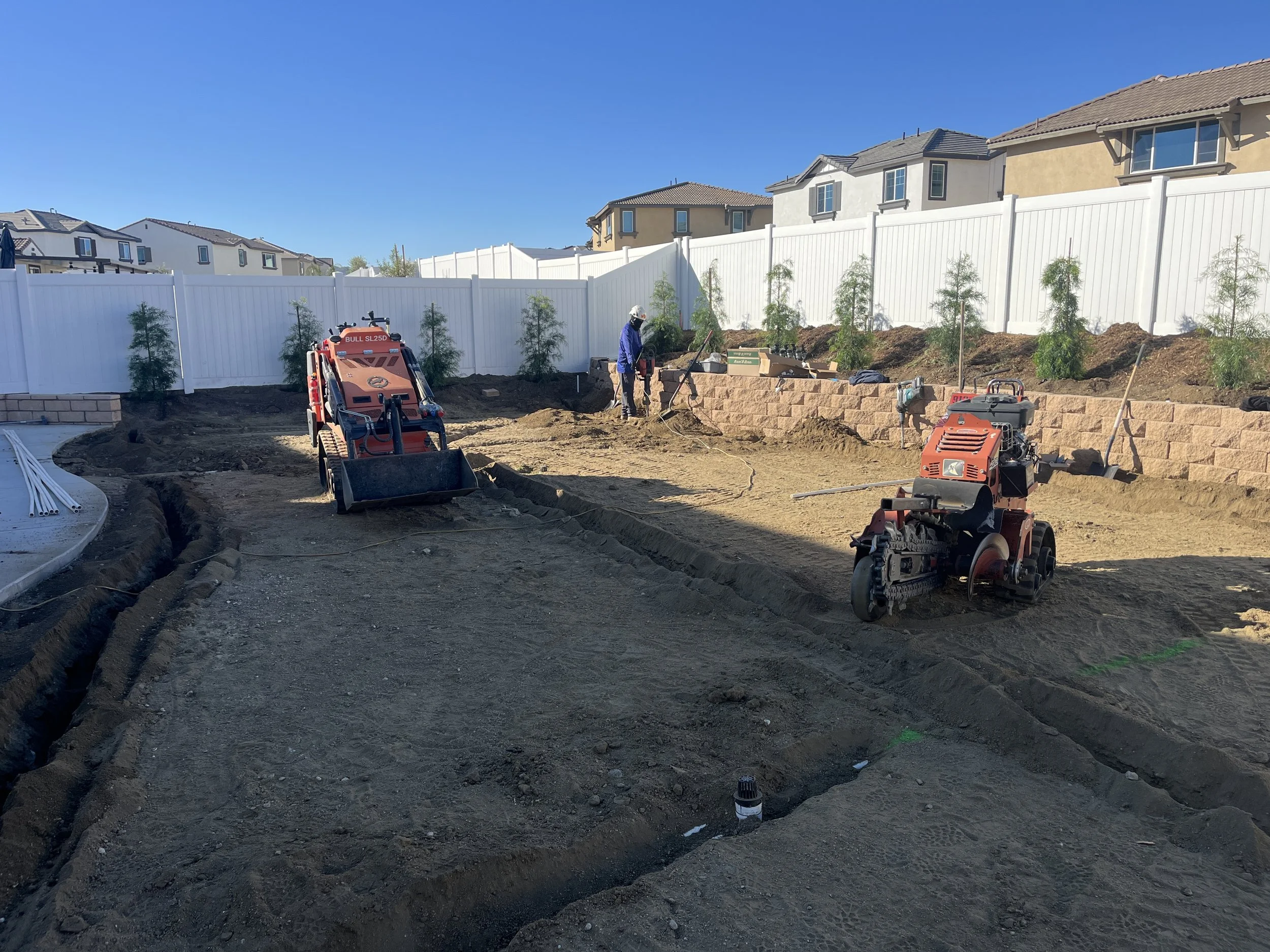 Construction site with two small compactors working on the dirt. A worker in a blue jacket and white hat is in the background, near a retaining wall of bricks. The area is surrounded by a white fence, and there are houses in the distance. It is a sun