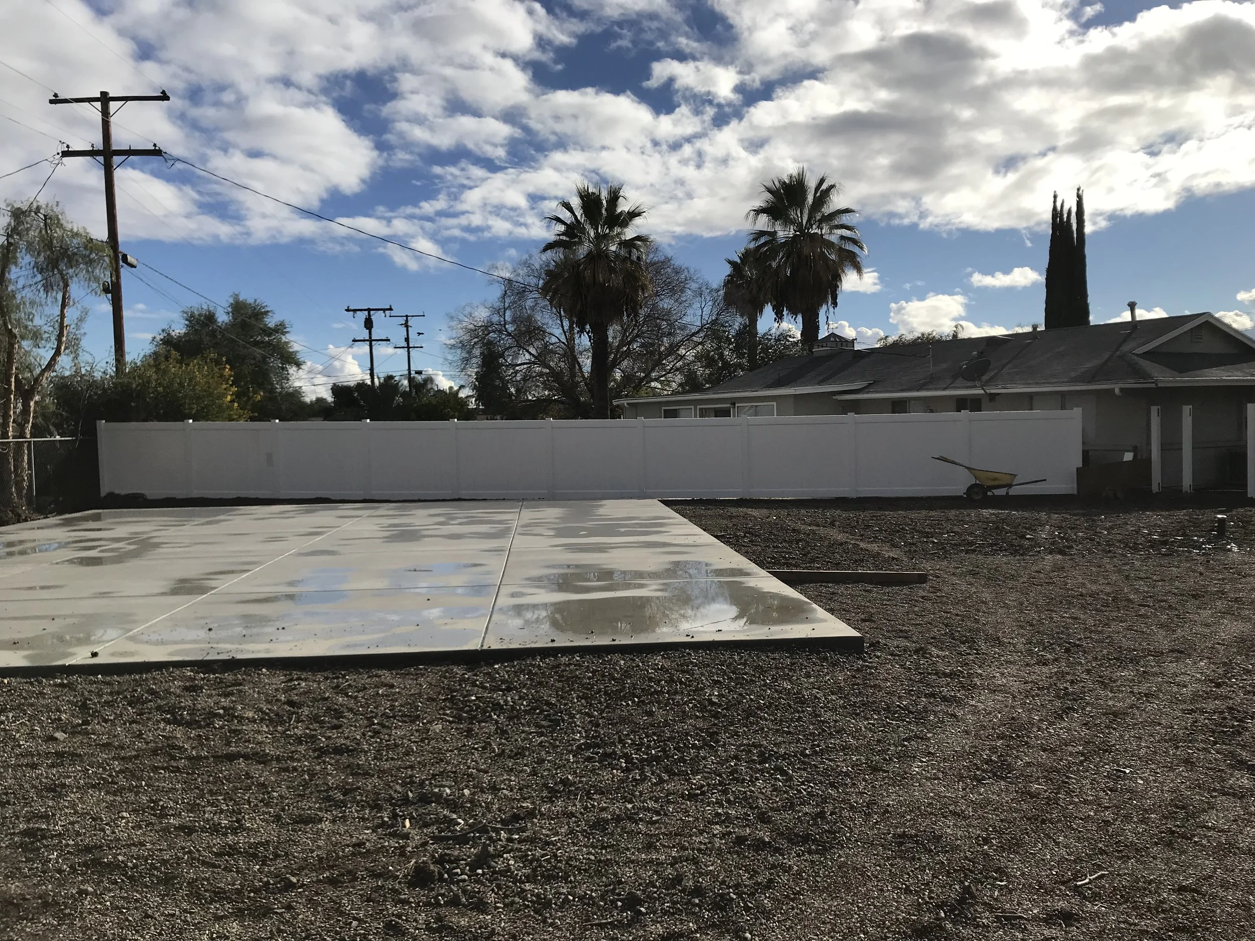 A backyard with a white fence, palm trees, a house, a gravel ground, a wheelbarrow, and a concrete pad under a partly cloudy sky.