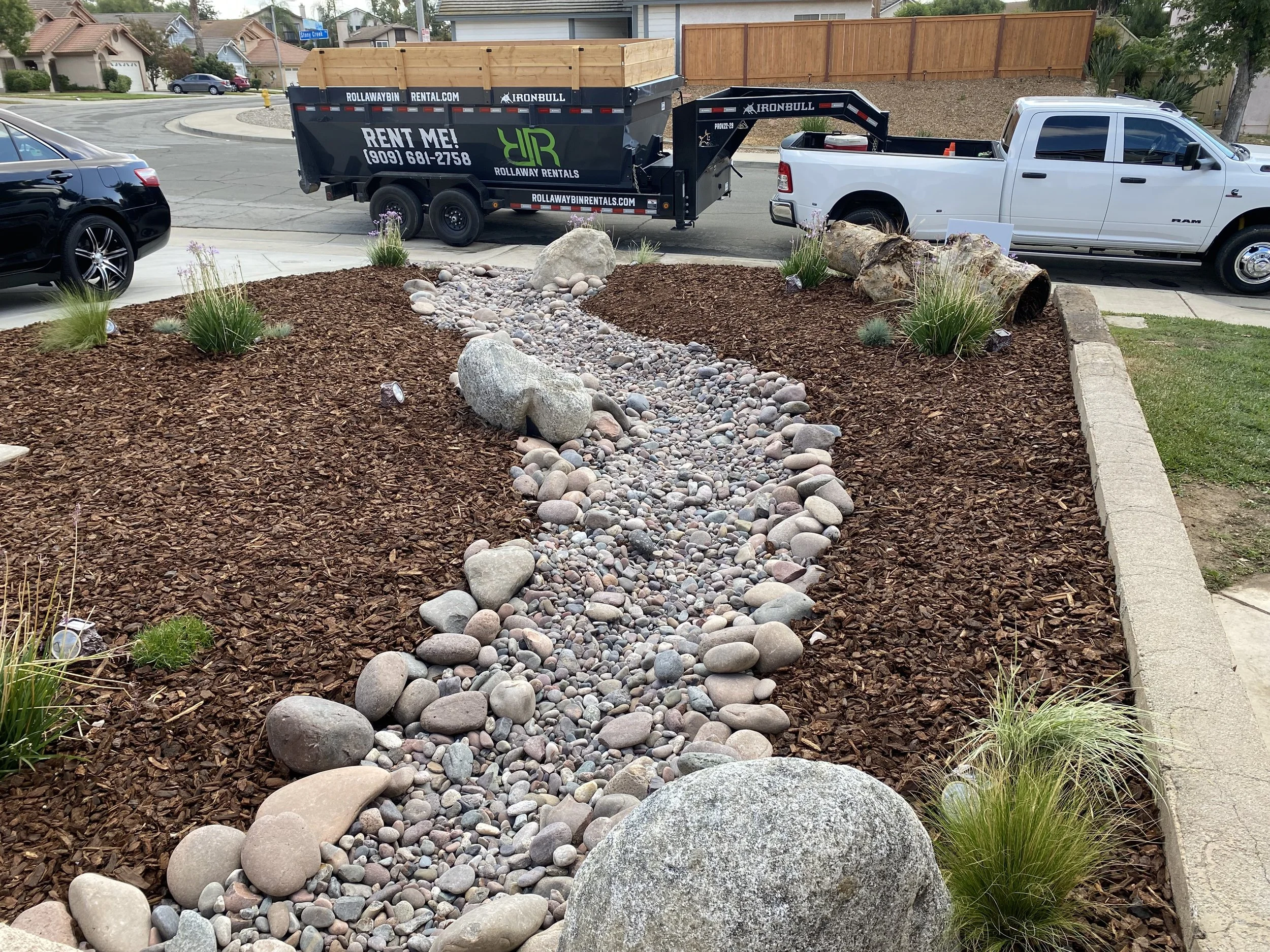 A landscaped yard with a dry creek bed made of various sizes of smooth, rounded stones and surrounded by brown mulch, with some green plants and rocks, next to a sidewalk and street with parked trucks and cars.