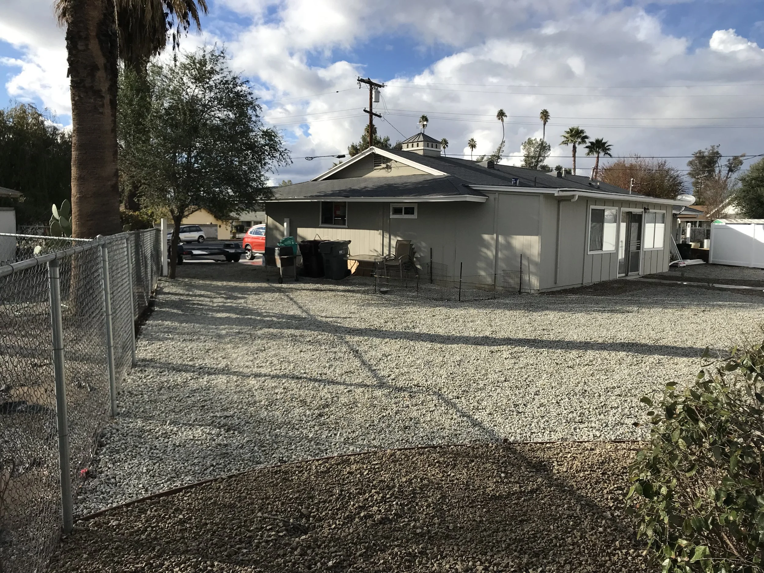A house with a gravel backyard, surrounded by chain link fencing, with palm trees and a partly cloudy sky in the background.