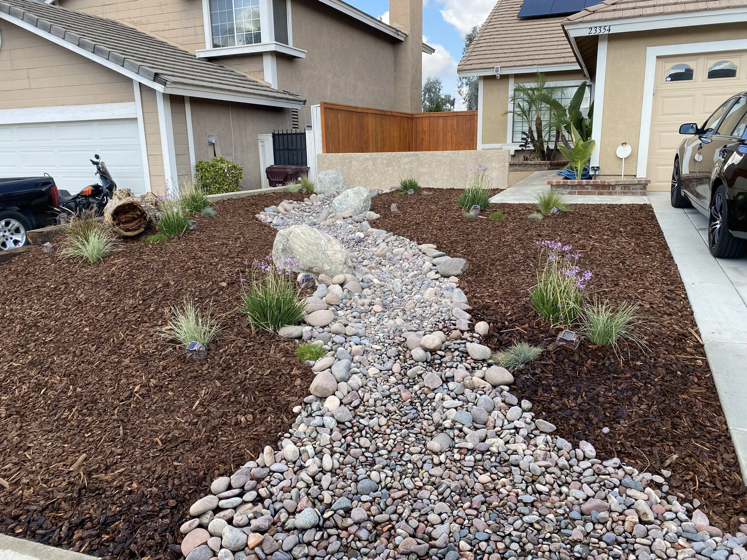 Front yard landscape with mulch, rocks, and plants in front of suburban houses, driveway, and parked cars.