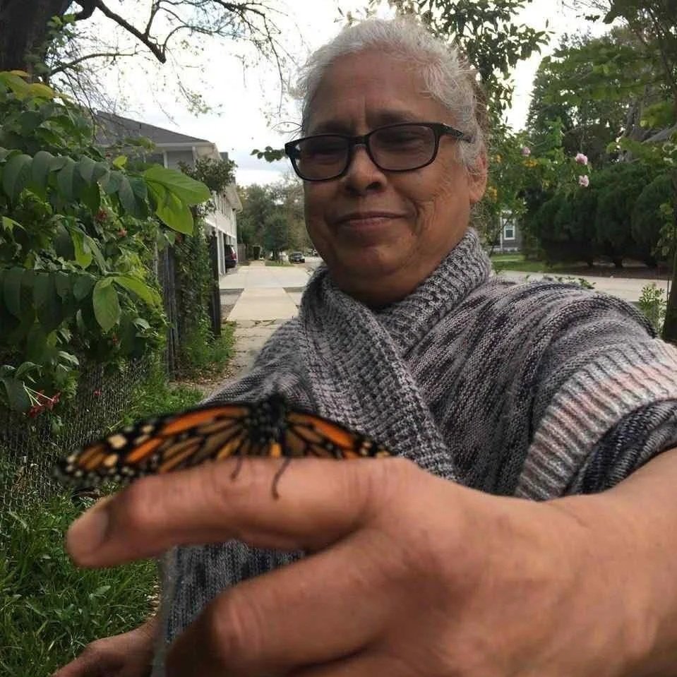 An elderly woman with glasses holding a monarch butterfly on her finger outdoors, smiling, with trees and houses in the background.