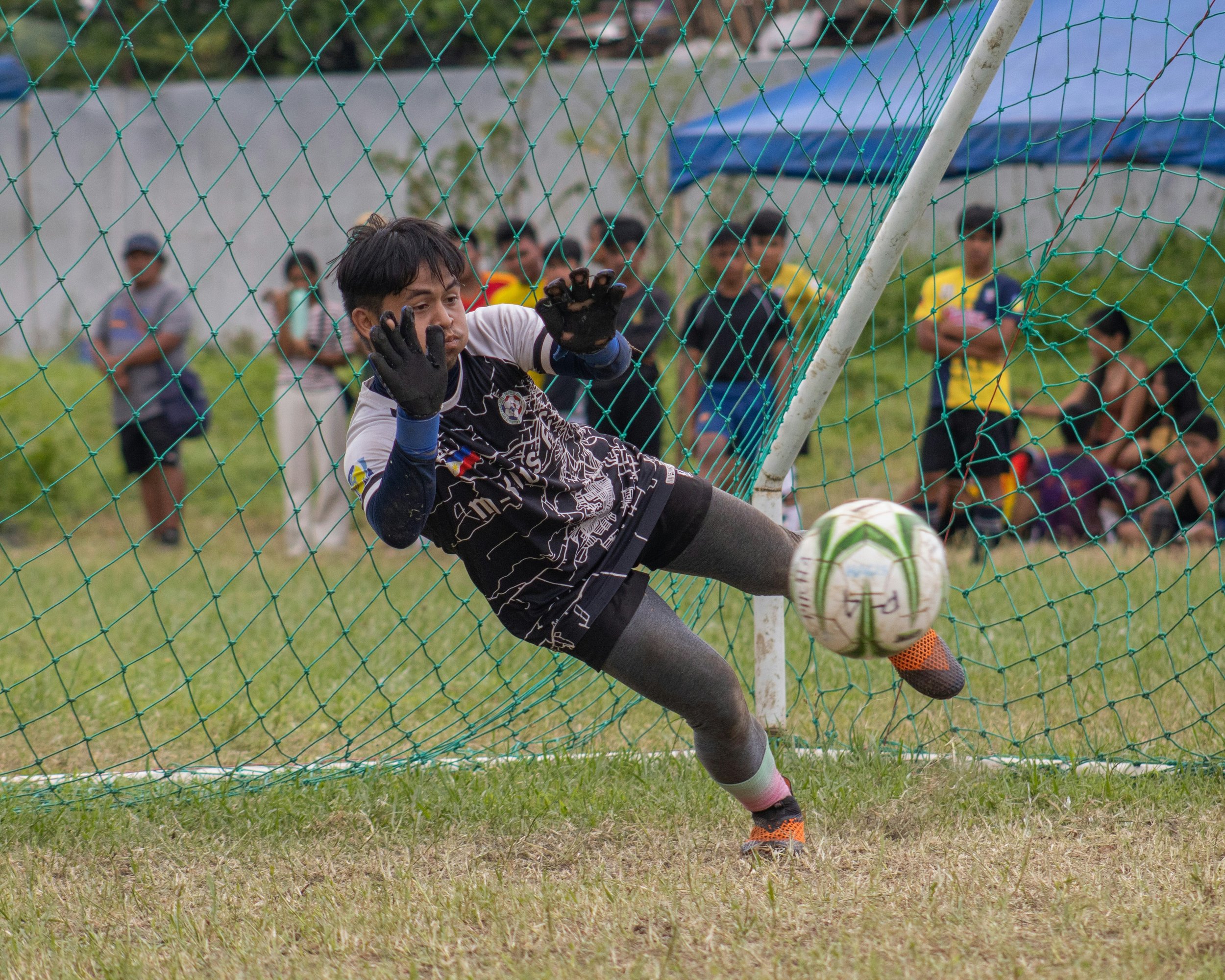 Soccer goalkeeper in black and white jersey diving to save a ball in the goal with green net, surrounded by onlookers on grass field.