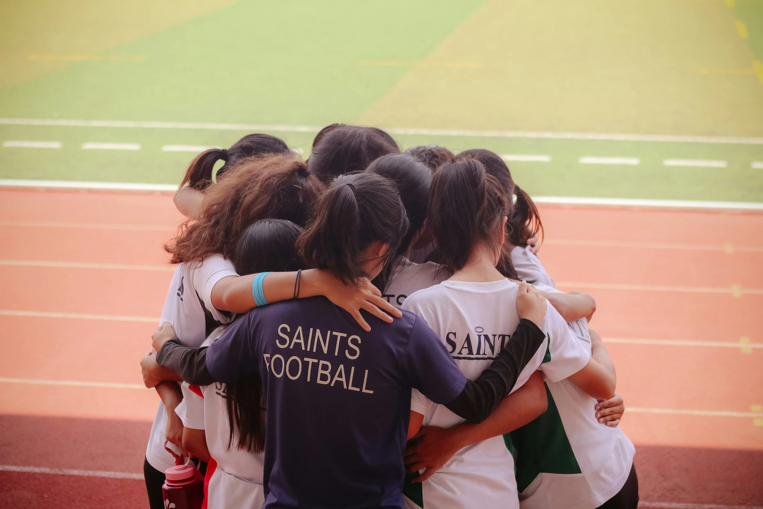 A group of women in sportswear hugging in a team huddle on a track field, with 