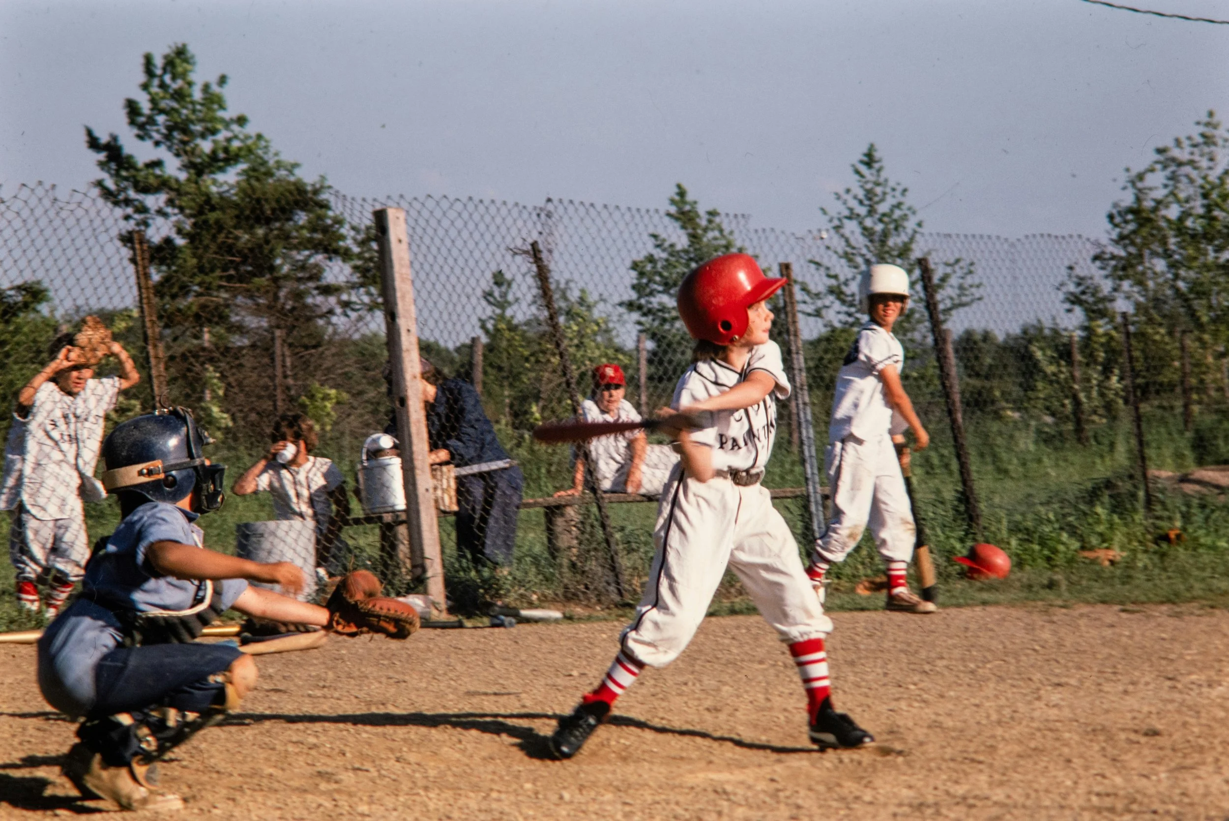 Children playing baseball in a baseball field, wearing uniforms with helmets and baseball gloves, with a chain-link fence and trees in the background.