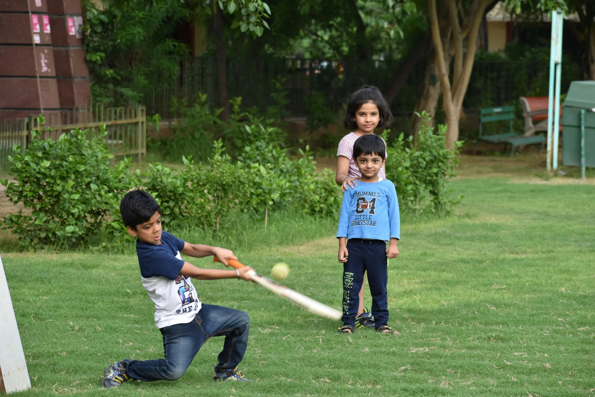 Three children playing cricket outdoors on a grassy lawn, with trees and benches in the background.