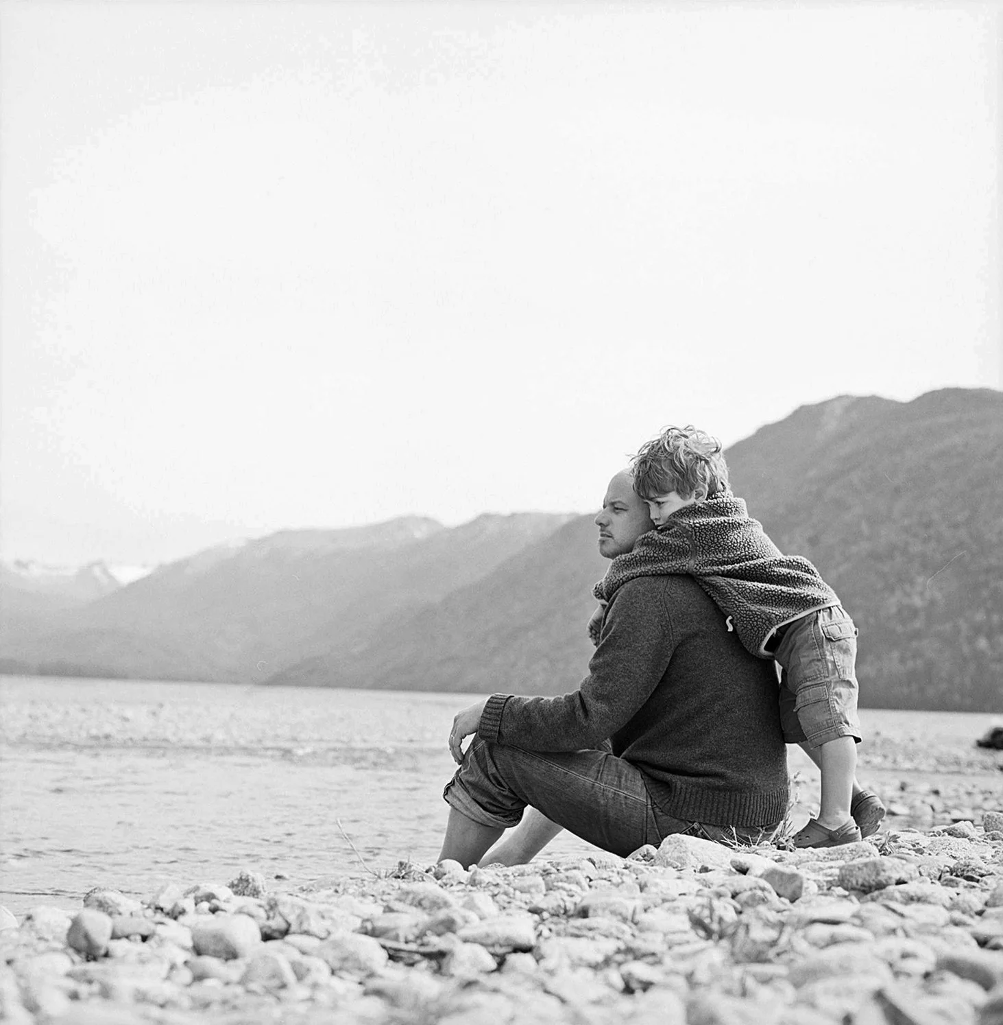 Black and white portrait of parent and child embracing on rocky beach with mountain backdrop.