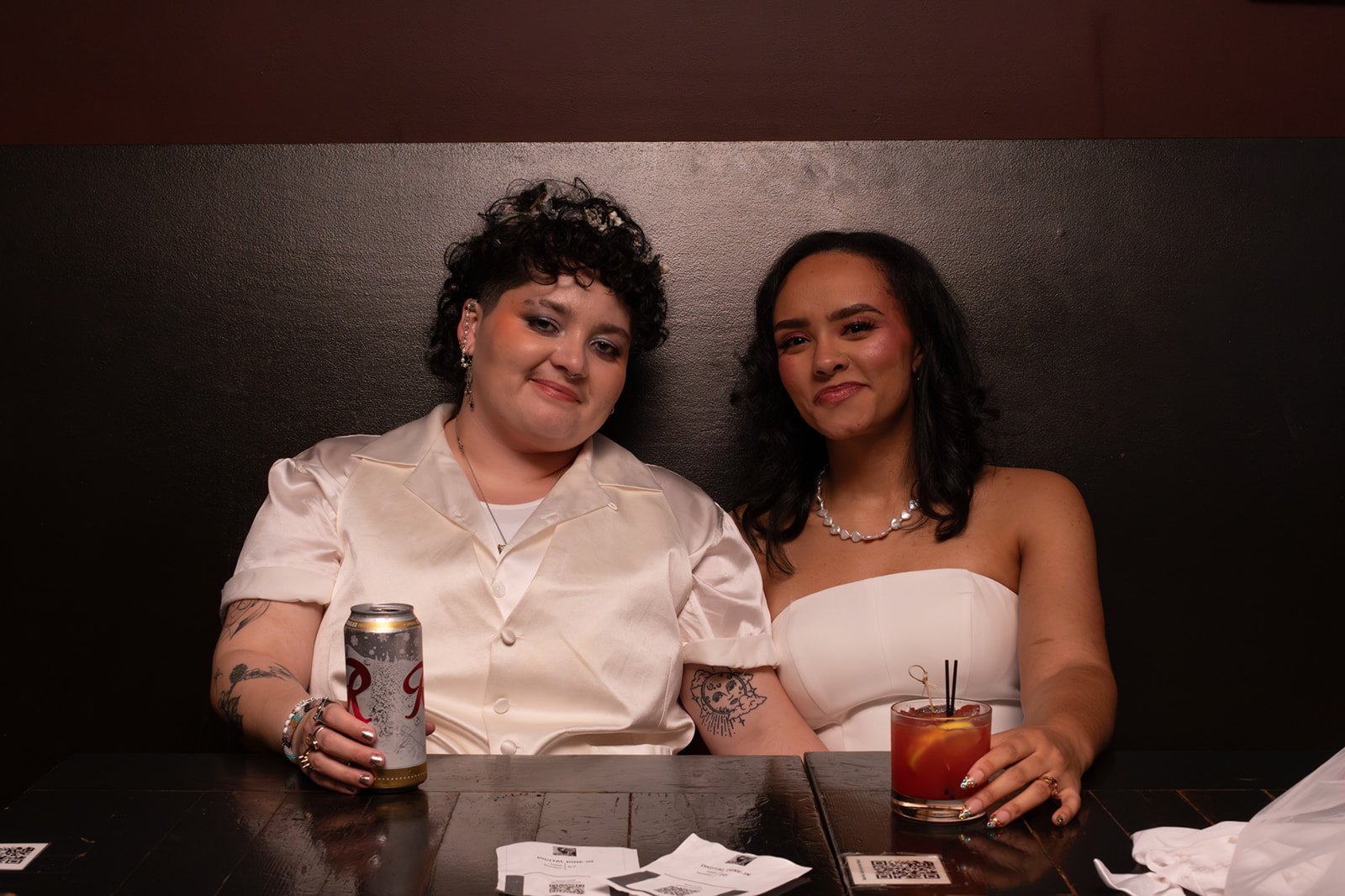 Bride and groom sitting together at their wedding reception, relaxed and smiling while sharing drinks at a wooden table.