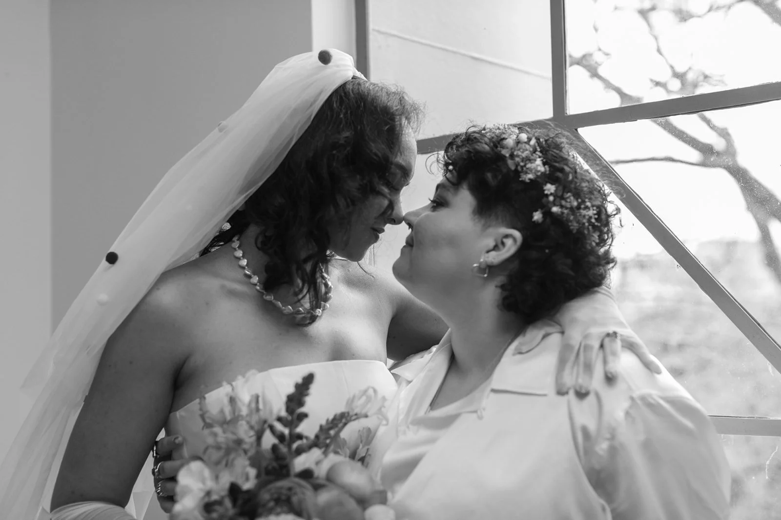 Black and white portrait of two brides sharing a quiet, intimate moment by a window on their wedding day.