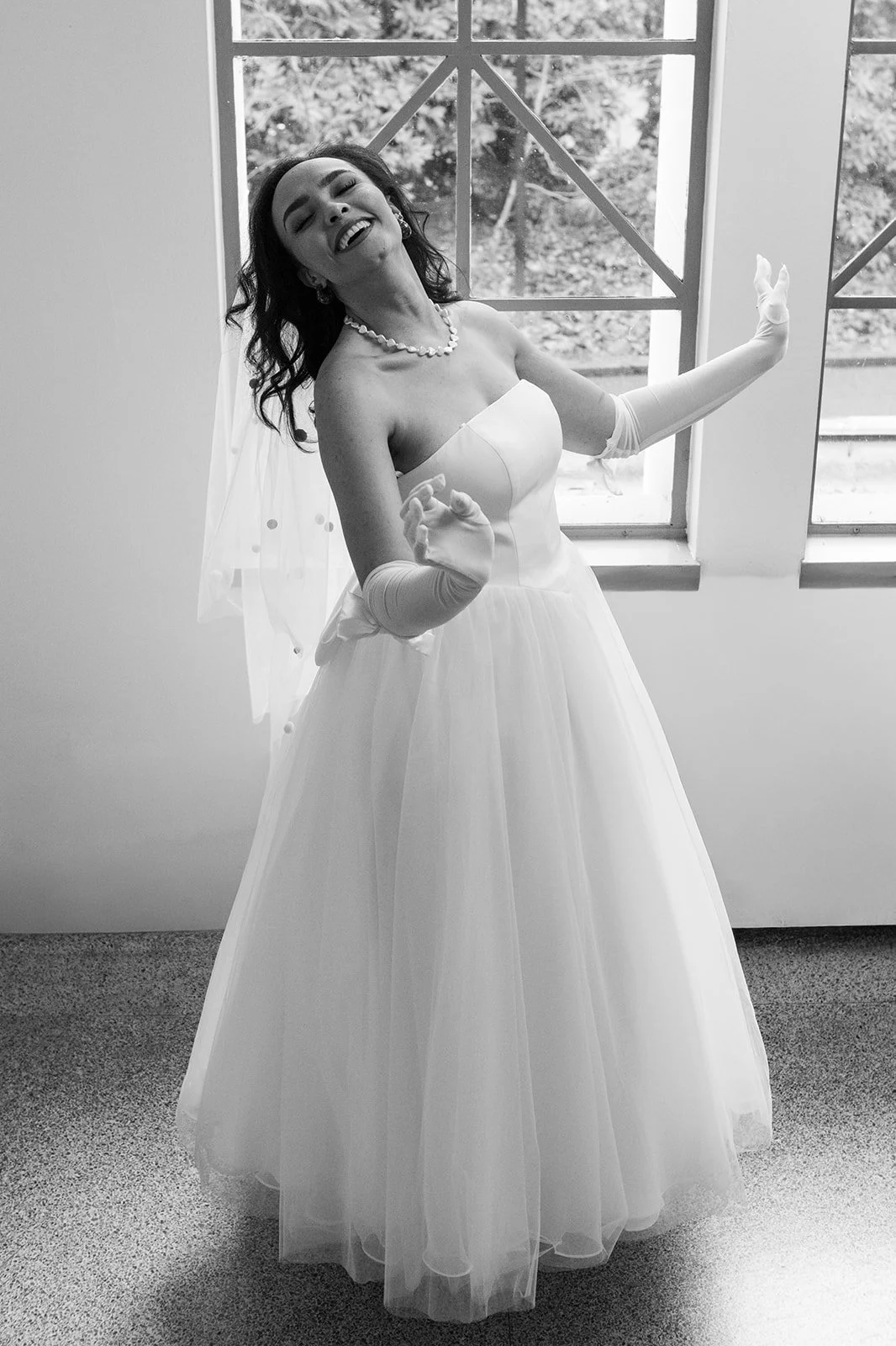 Black and white portrait of the bride dancing joyfully in her wedding dress beside a large window.