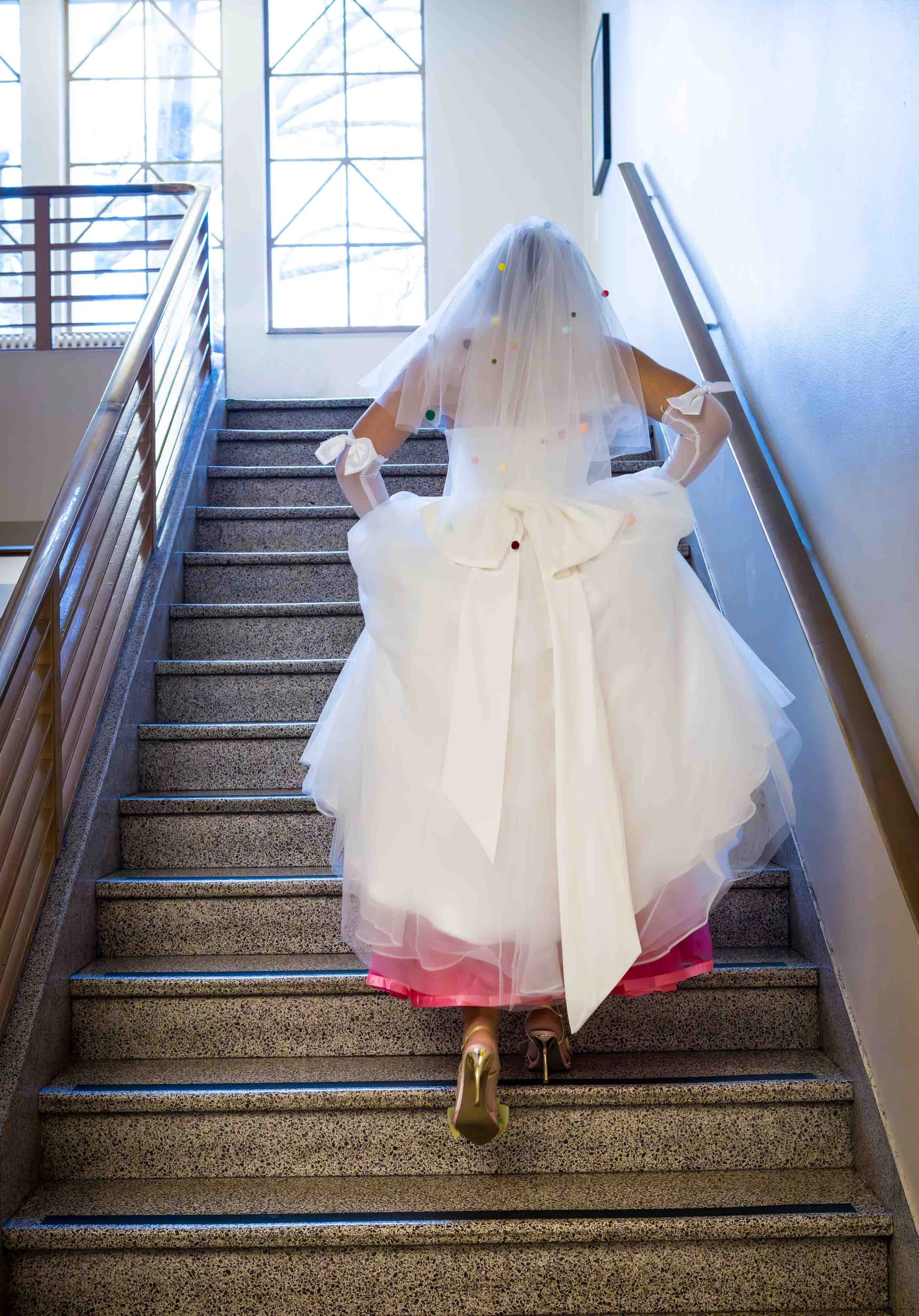 Bride walking upstairs in her wedding dress, veil and layered pink tulle catching the light in a quiet, anticipatory moment.