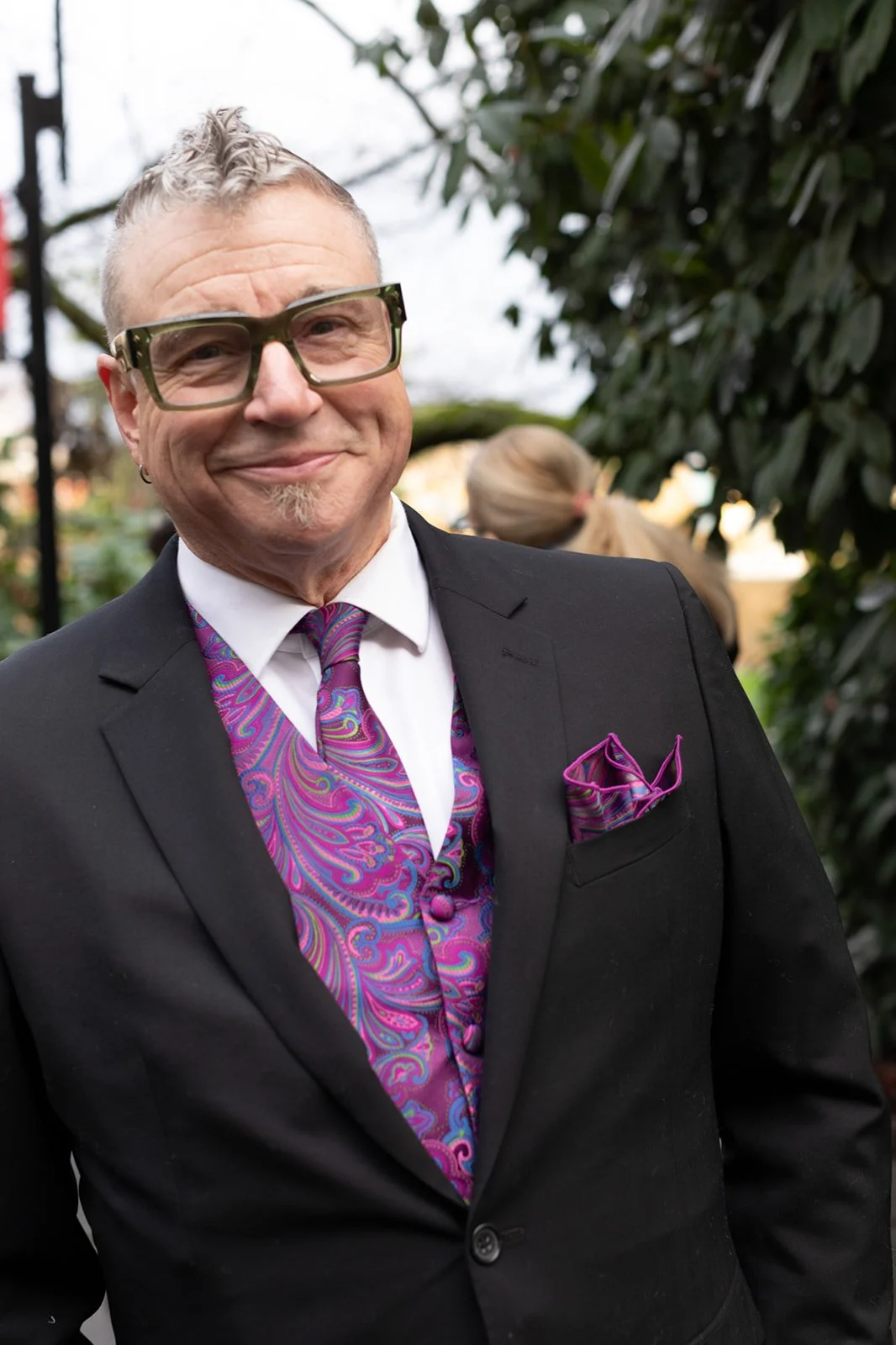 Wedding guest smiling in a tailored suit with colorful vest and tie, photographed outdoors during the celebration.