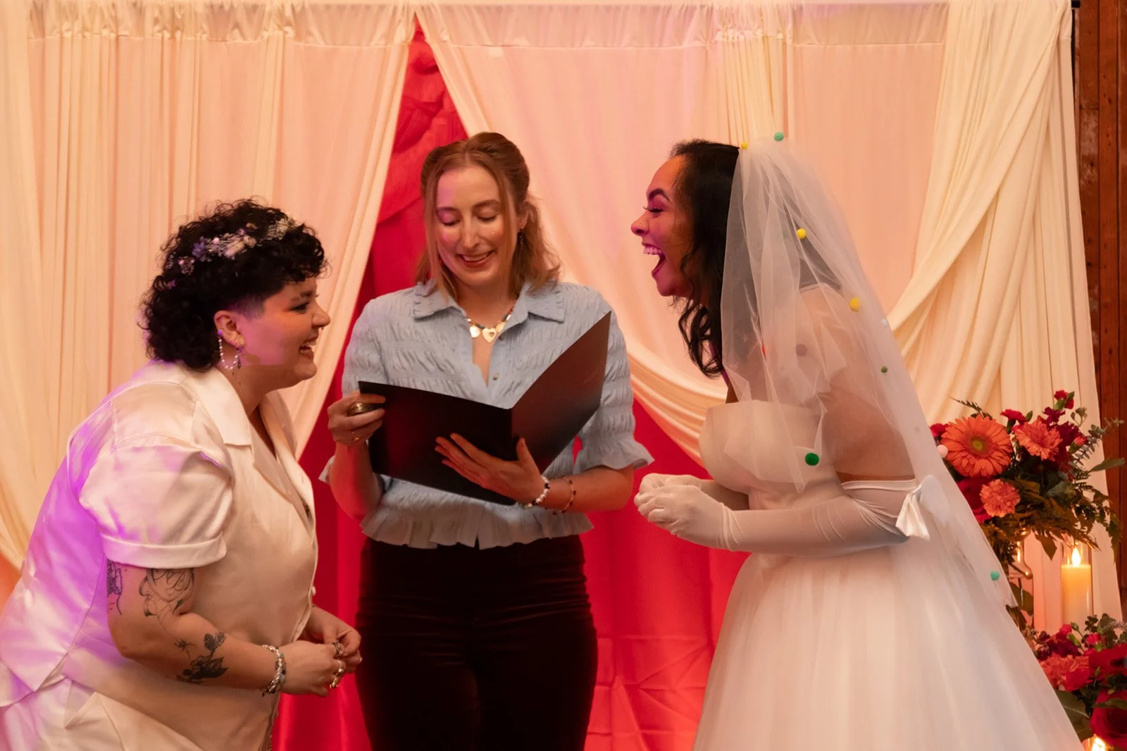 Queer wedding ceremony as an officiant reads vows while the bride and groom laugh beneath a colorful backdrop.