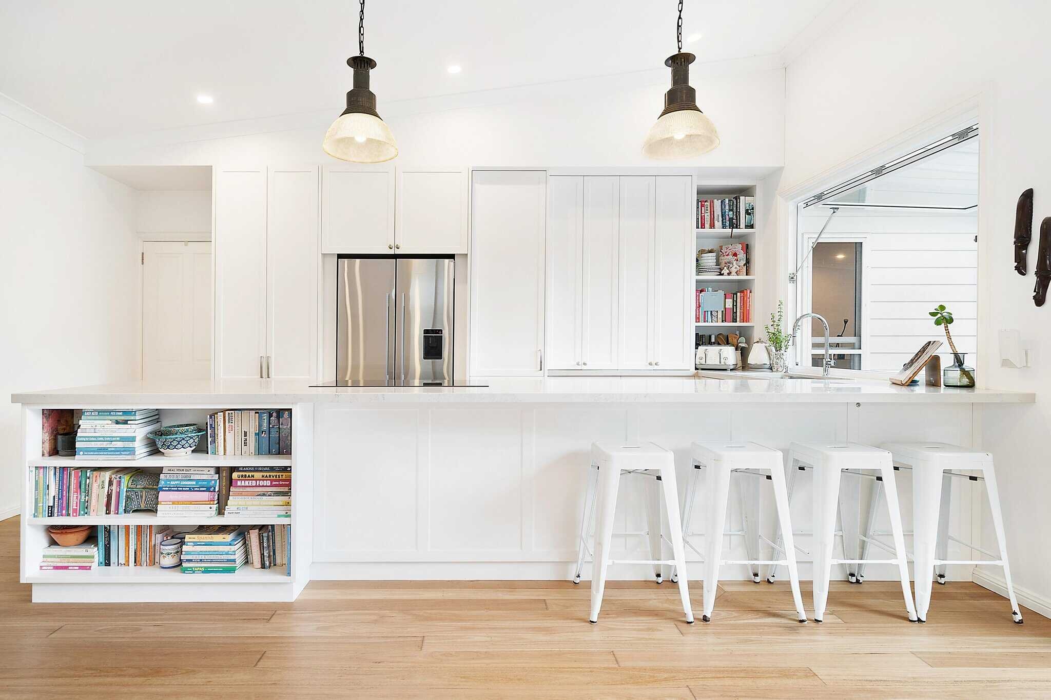 Modern white kitchen with bar stools, pendant lights, wood flooring, built-in bookshelves, stainless steel refrigerator, and a window view.