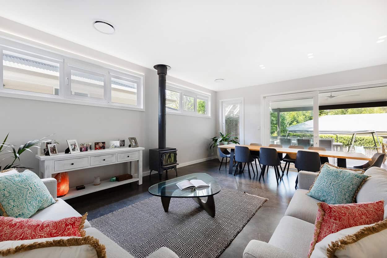 Modern living and dining area with a white console table displaying photographs, a wood-burning stove, glass coffee table, sofas with colorful cushions, and a long dining table with dark chairs next to large glass doors opening to a patio.