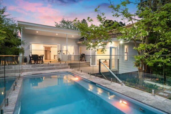 Modern house with a pool in the backyard, surrounded by glass fencing and greenery, under a blue and pink evening sky.