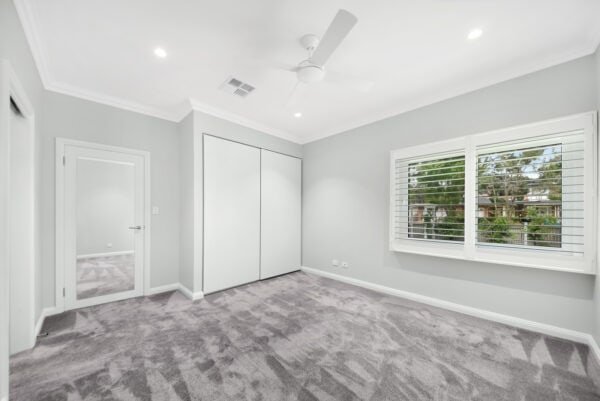 Empty bedroom with gray carpet, white walls, ceiling fan, built-in wardrobe, large window with white shutters, and a full-length mirror on the door.