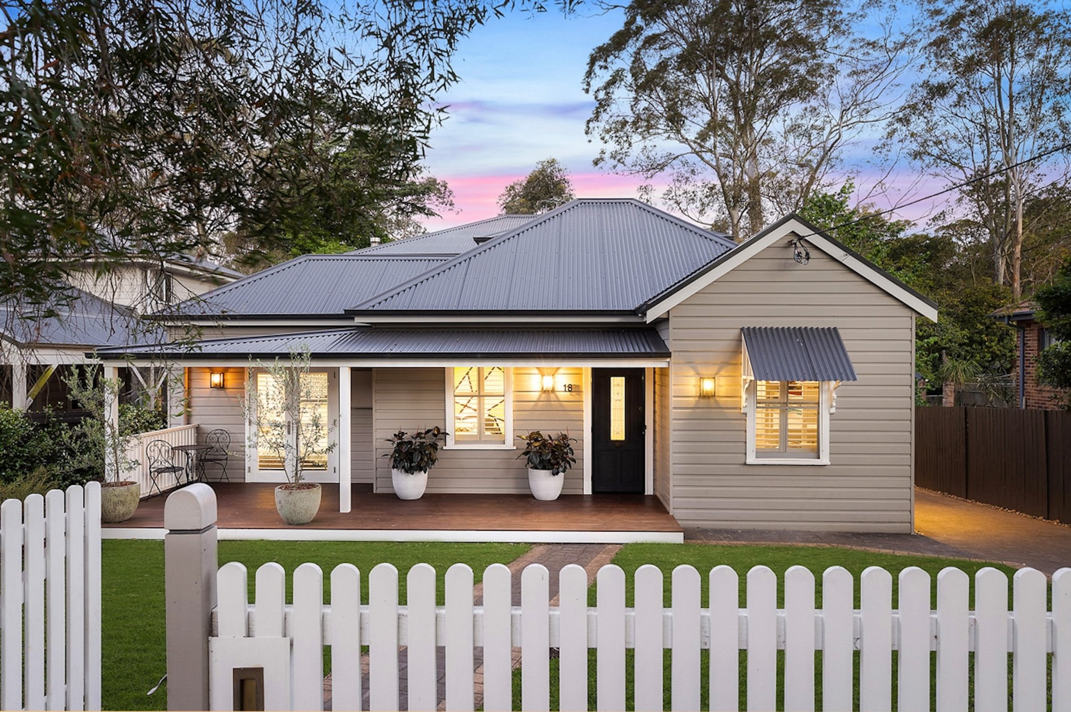 Charming single-story house with a gray metal roof, beige siding, and a welcoming front porch, surrounded by a white picket fence and garden, set against a twilight sky.