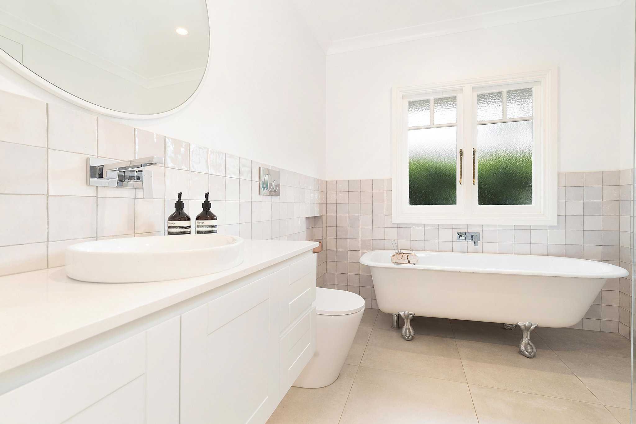 Modern bathroom with white freestanding bathtub, white vanity with oval sink, wall-mounted faucet, two soap dispensers, round mirror, tiled walls, and frosted window.