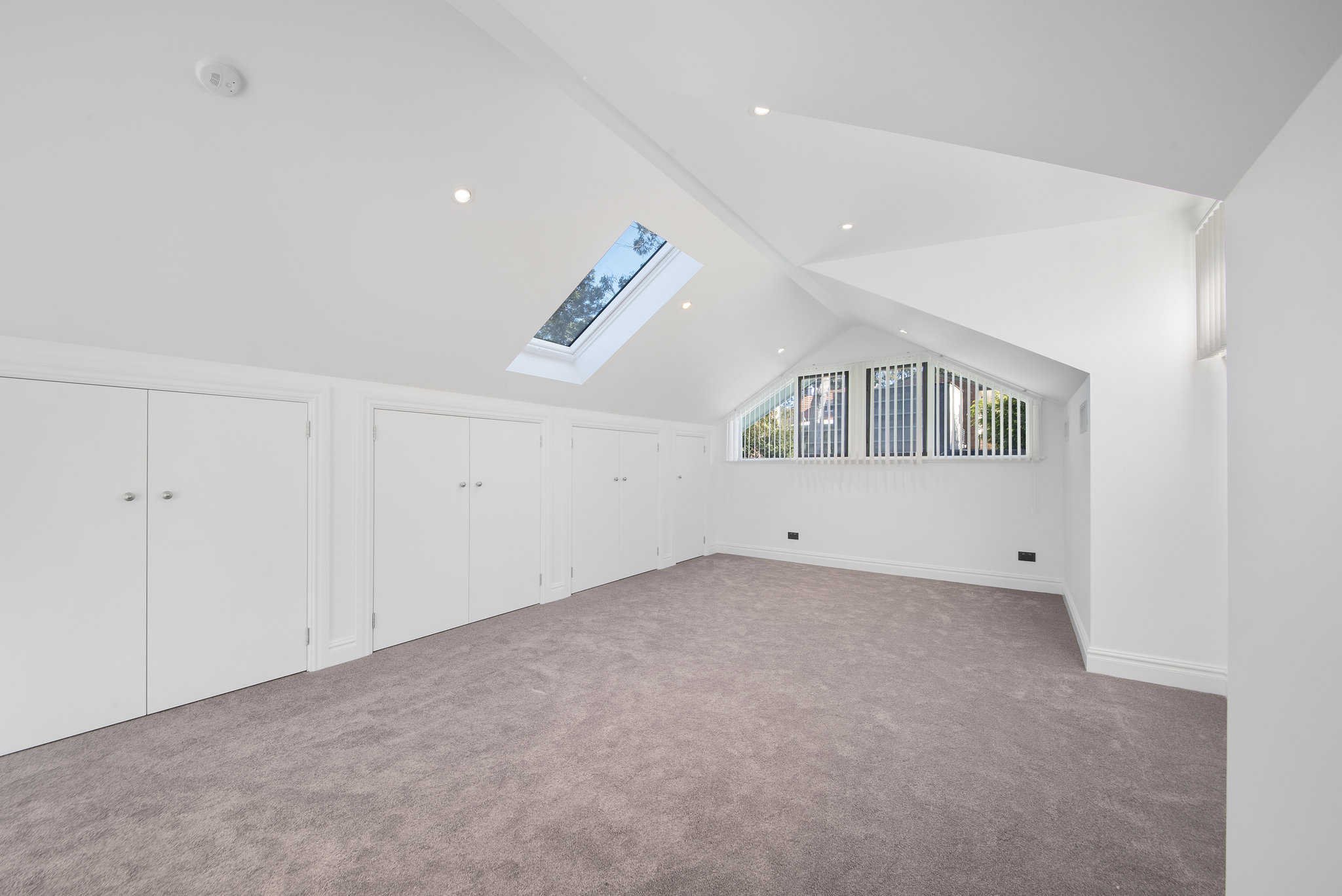 Spacious attic room with white walls, carpeted floor, large triangular window, ceiling skylight, and built-in storage cabinets.