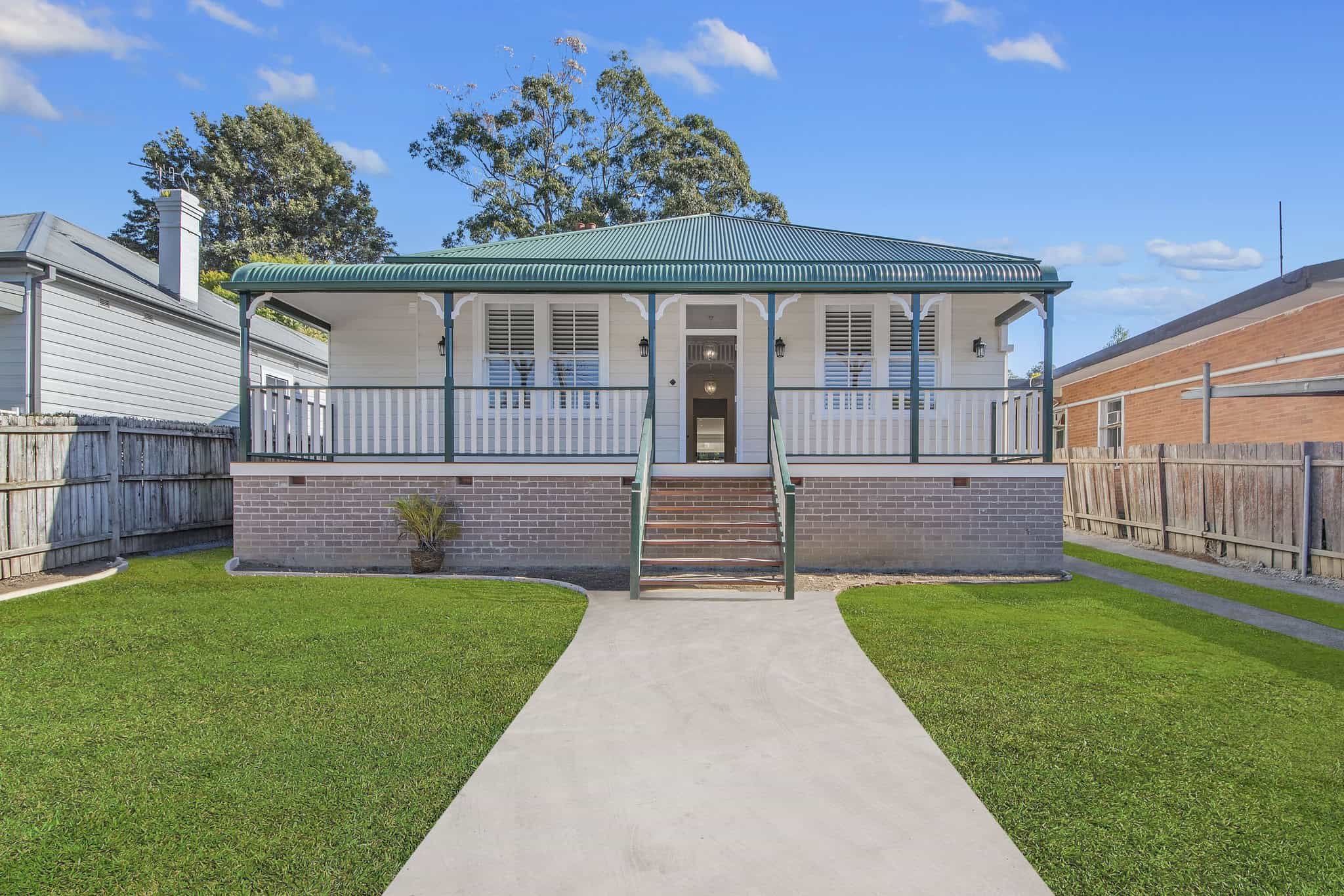Front view of a single-story house with a green metal roof, white facade, and a wraparound porch. The house is elevated with stairs leading to the entrance and surrounded by a neatly maintained lawn. A wooden fence lines the side of the property.