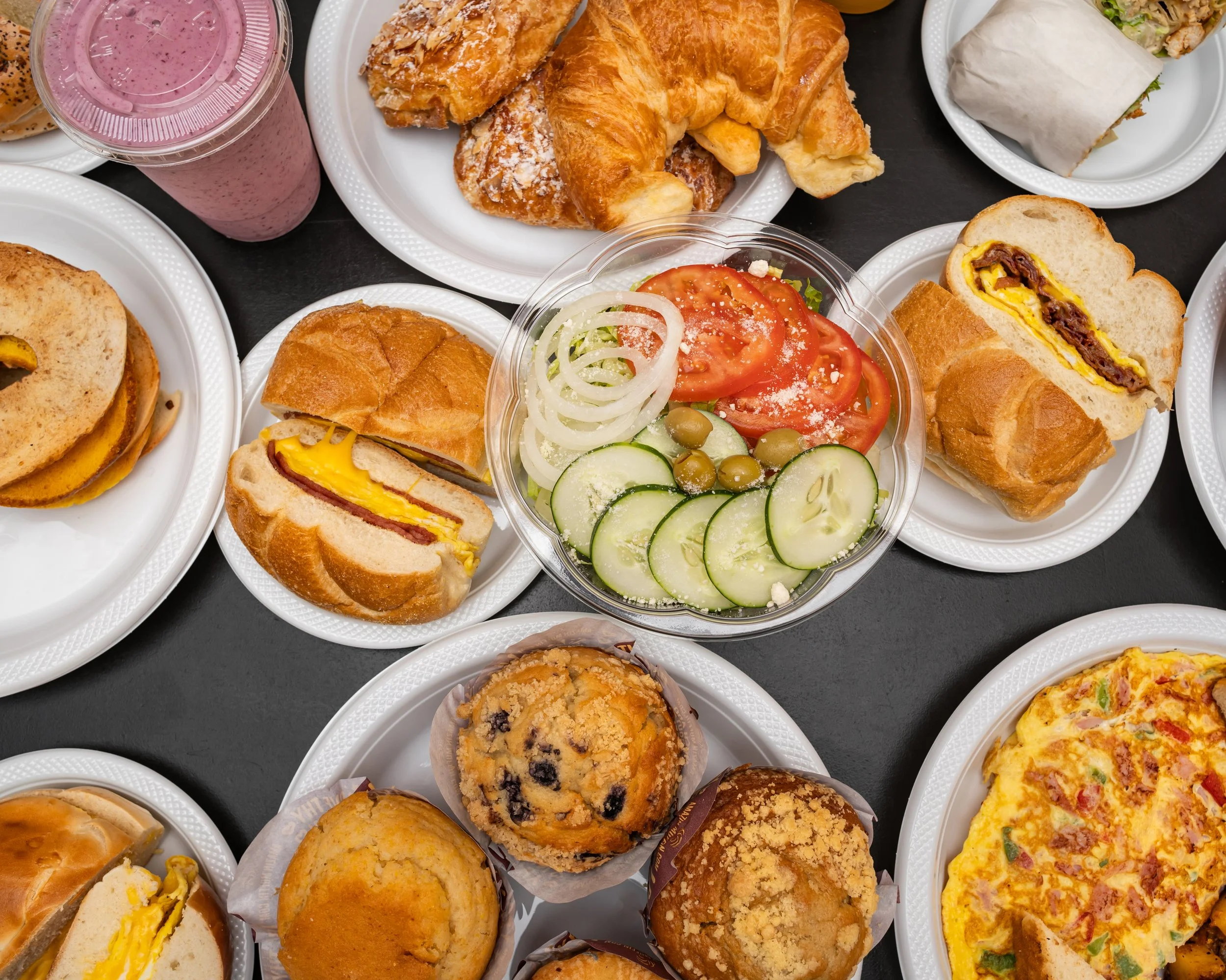 Assorted breakfast foods and snacks including croissants, sandwiches, salads, muffins, cupcakes, and smoothies on a table.