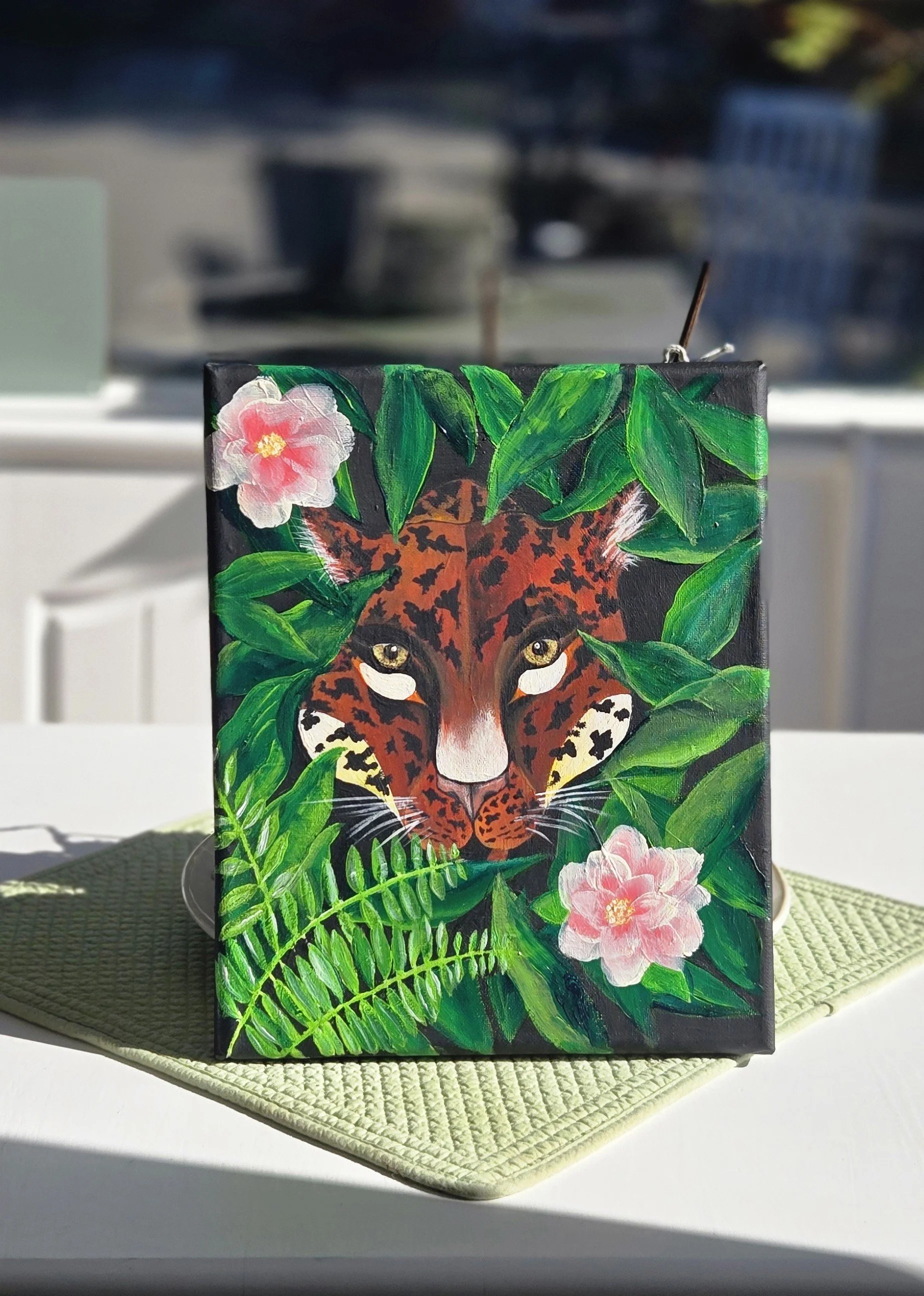 A painting of a leopard’s face surrounded by green leaves and pink flowers, displayed on a black canvas.