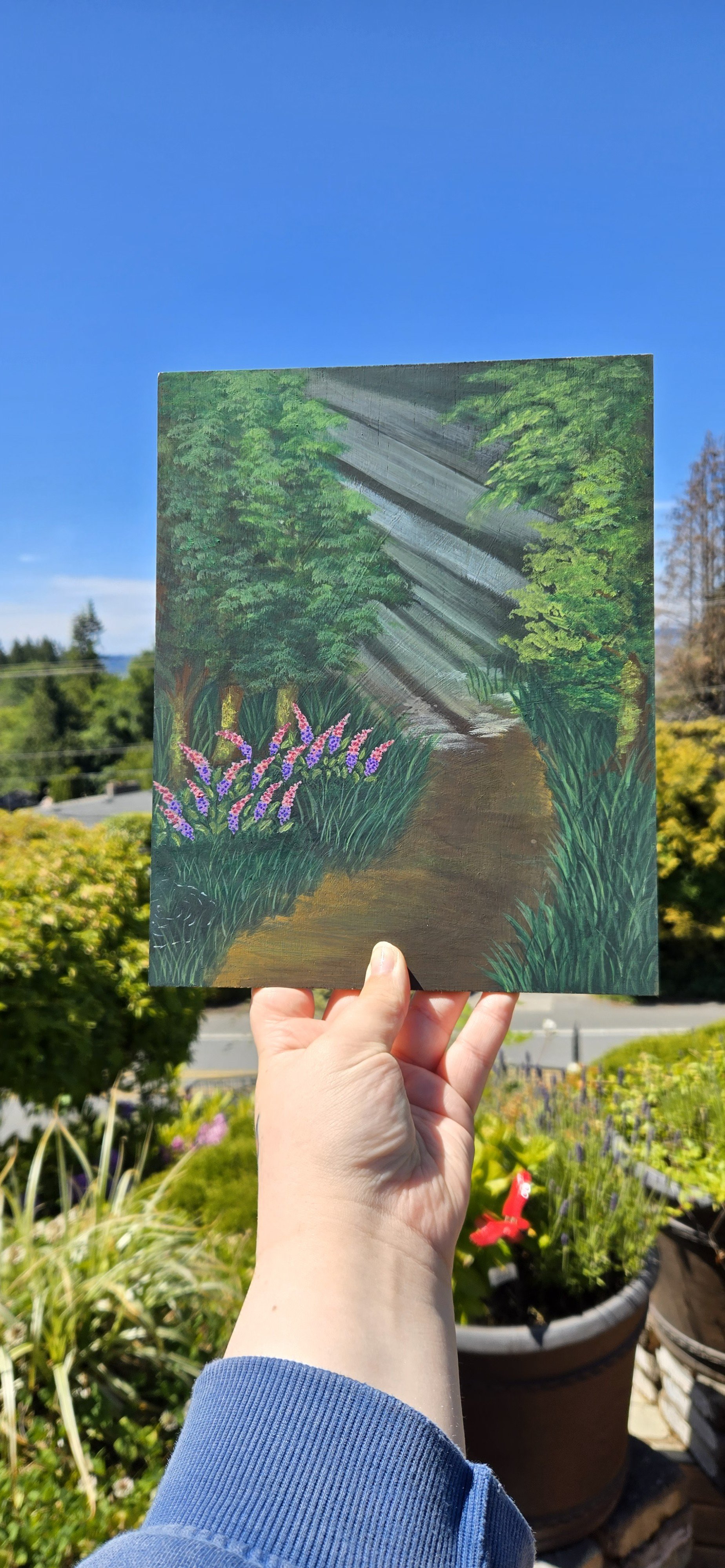Hand holding a painted board with a forest scene, pathway, trees, and purple flowers, with outdoor garden and clear blue sky in the background.