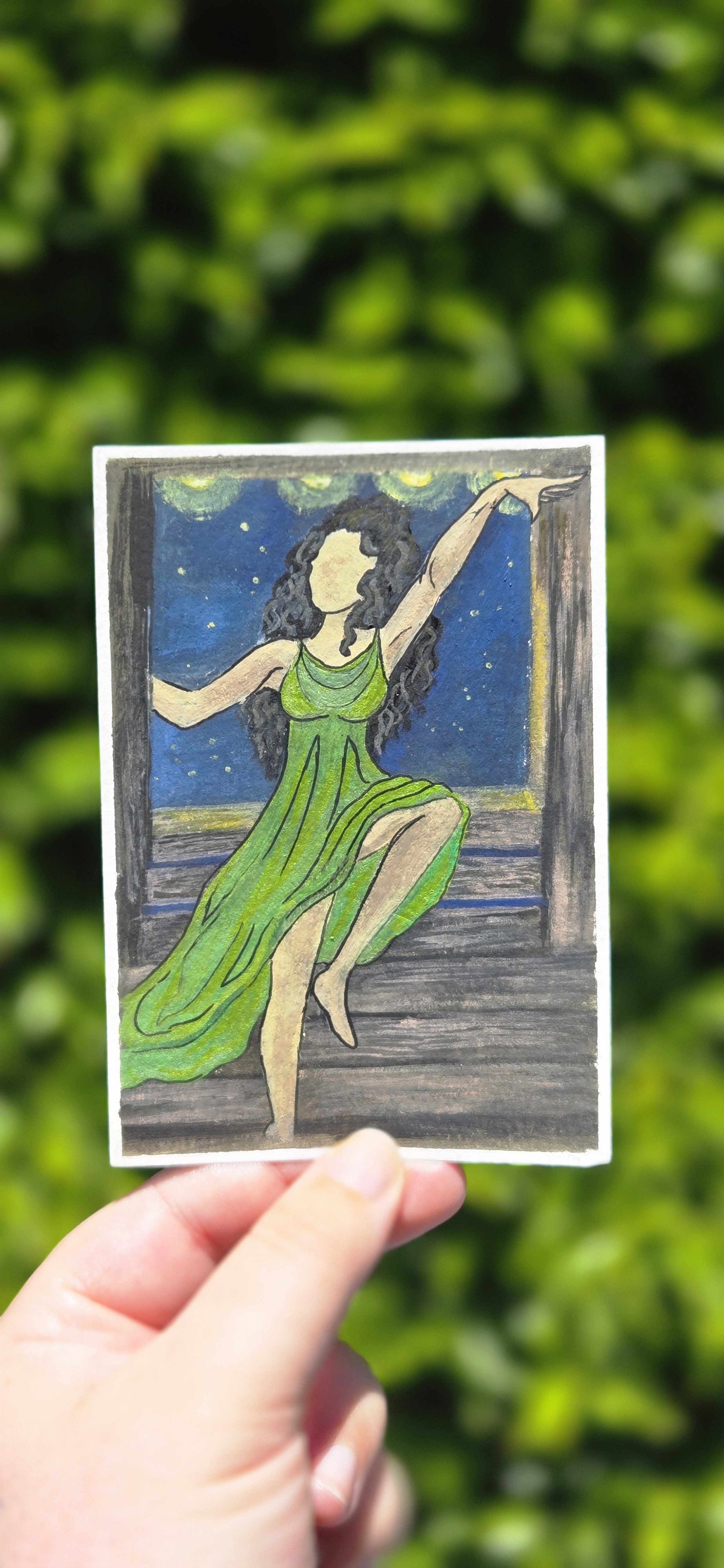Hand holding a colored drawing of a woman in a green dress sitting by a window, with a night sky and stars visible outside.