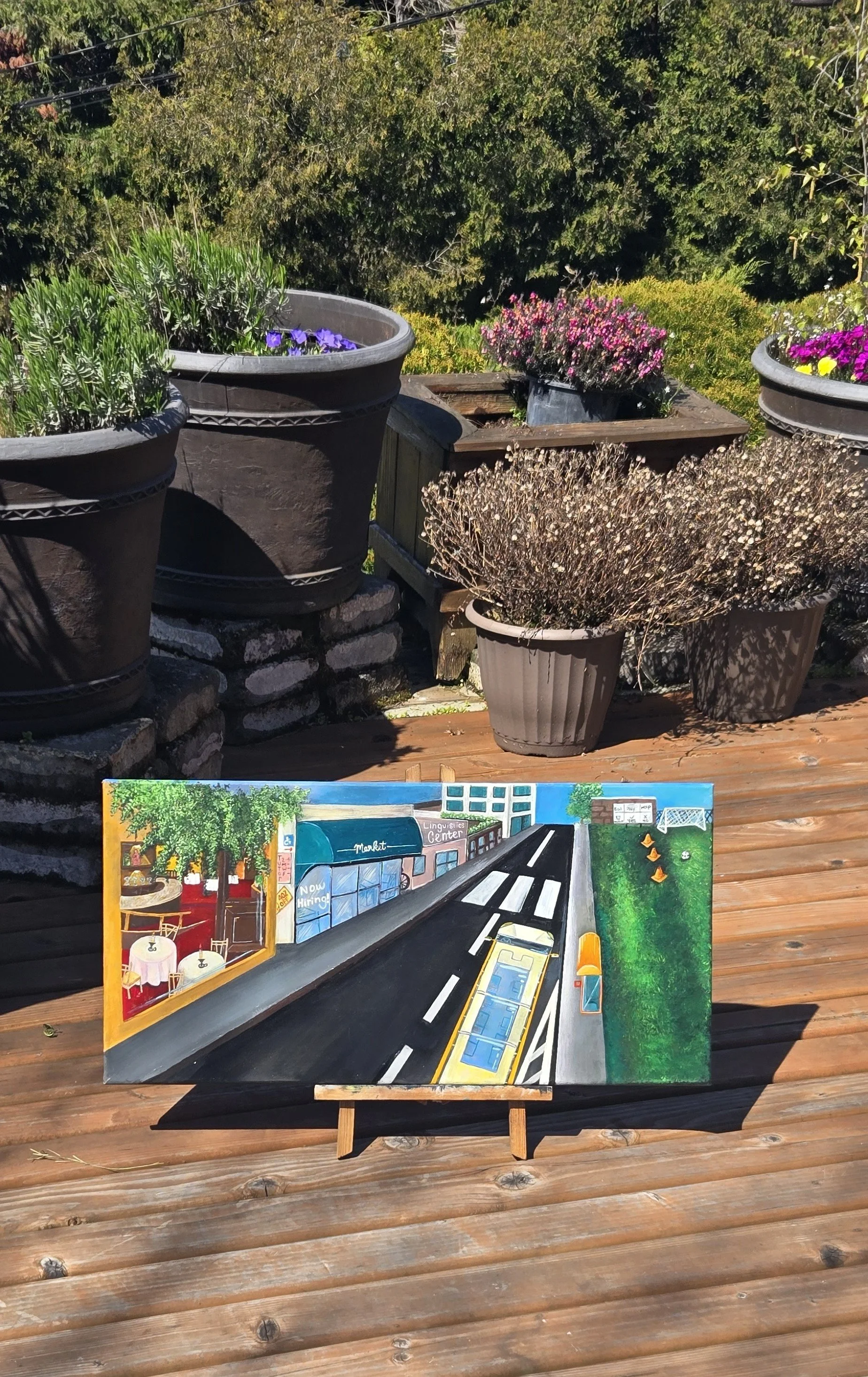 A colorful painting of a street scene with a bus stop and buildings, placed on an easel outdoors on a wooden deck, with potted plants and trees in the background.