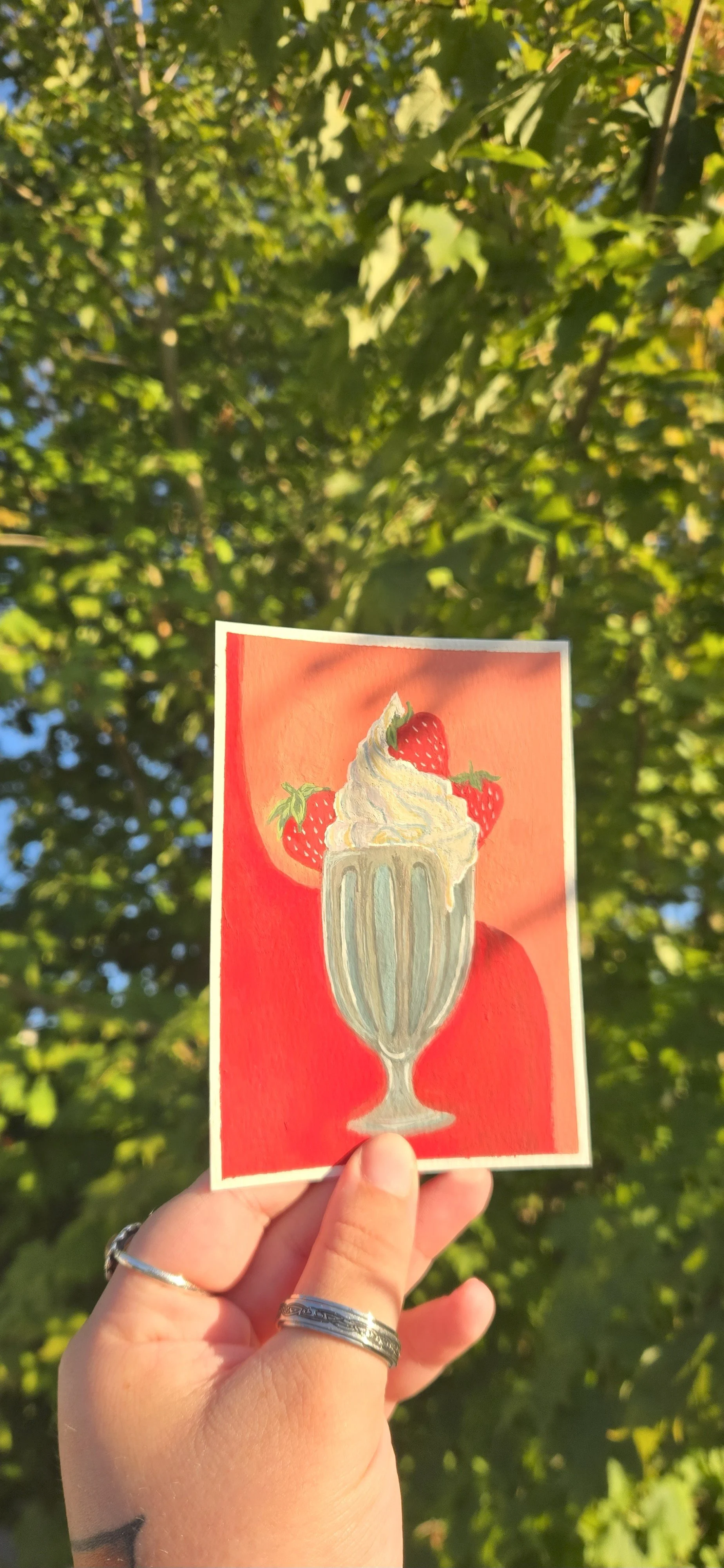A hand holding a colorful illustration of a strawberry sundae in a tall glass with whipped cream and strawberries, against a background of green leaves and blue sky.