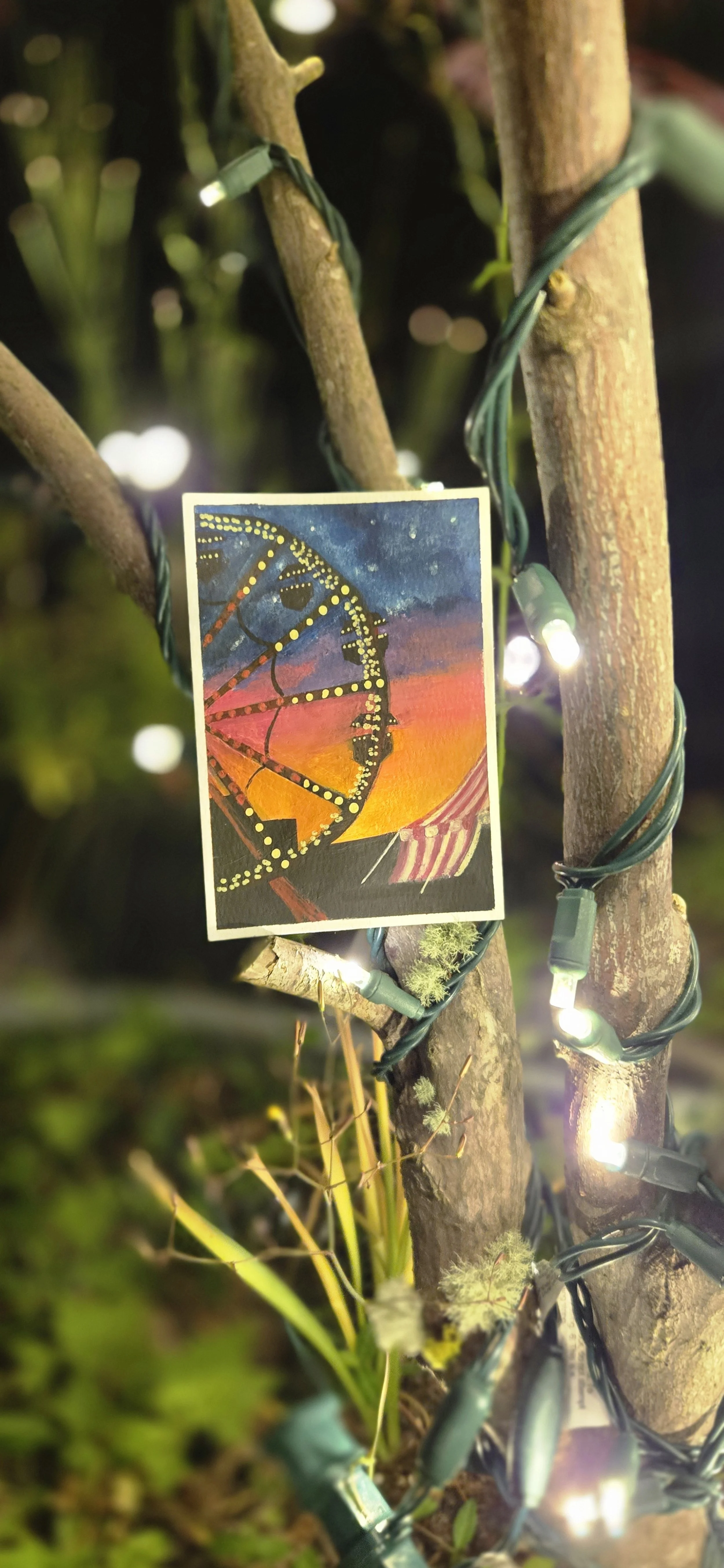 Close-up of a small card with a colorful sunset scene featuring a ferris wheel and striped tent, attached to two tree branches with string lights wrapped around them.